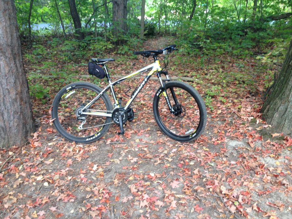 A mountain bike resting on the ground covered with fallen leaves, surrounded by trees in a wooded area. Maybury mountain bike trail.