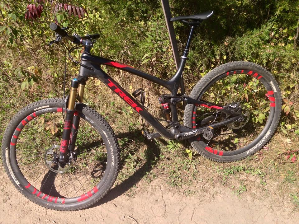 Trek Fuel EX 9.9 29 XTR: A black and red Trek mountain bike is parked on a dirt path surrounded by greenery. The bike features thick tires, a sporty frame, and suspension components, indicating it's designed for off-road riding. Sunlight casts a warm glow on the scene, highlighting the bike's details against the natural backdrop.