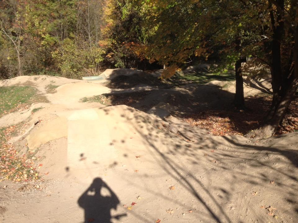 A dirt biking track in a wooded area, featuring several bumps and jumps. The scene captures autumn foliage, with vibrant yellow and orange leaves on the trees. A shadow of a person holding a camera is visible in the foreground. Novi Tree Farm (Lakeshore Park) mountain bike trail.
