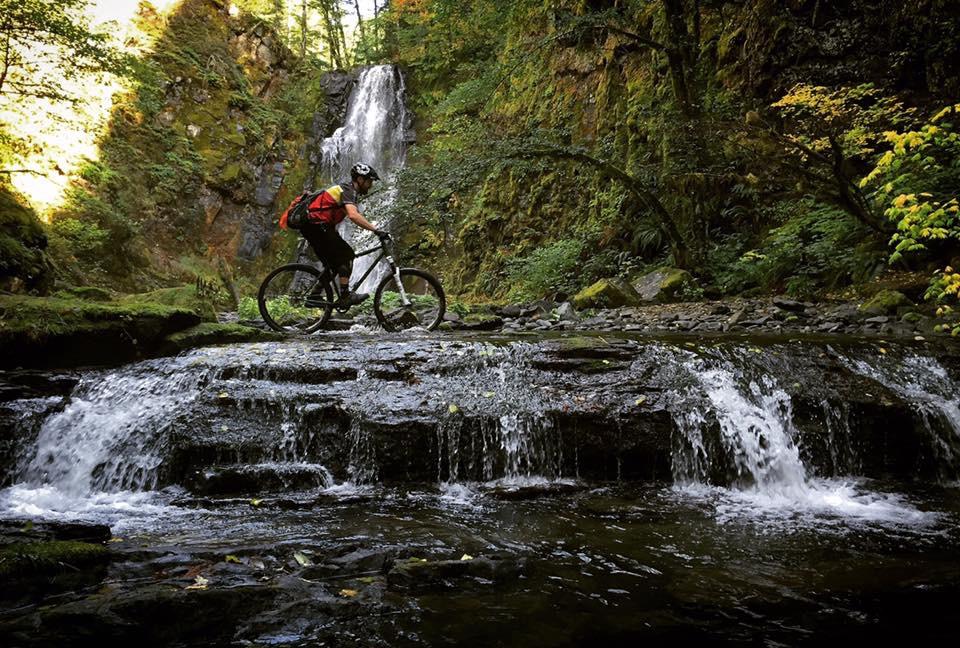 A mountain biker navigates a rocky stream bed near a waterfall, surrounded by lush green vegetation and moss-covered cliffs. The scene captures the dynamic interaction between outdoor adventure and nature. Siouxon Creek mountain bike trail.