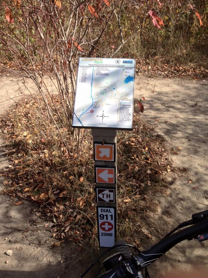 A trail map sign at a park, showing various paths and trail options, with directional arrows for navigation and a phone number to dial in case of emergencies. The surrounding area features brown foliage and a dirt path. A bicycle wheel is partially visible at the bottom right of the image. Novi Tree Farm (Lakeshore Park) mountain bike trail.