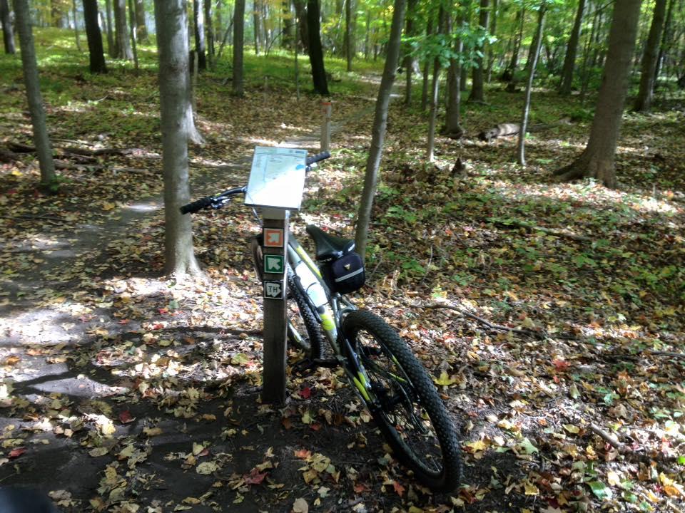 A mountain bike is leaned against a trail sign in a wooded area, surrounded by autumn leaves on the ground. The scene features tall trees with green and orange foliage, suggesting a serene outdoor setting ideal for cycling and exploring nature. Novi Tree Farm (Lakeshore Park) mountain bike trail.