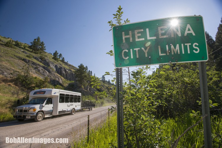 A green sign reading "Helena City Limits" is prominently displayed on the side of a road. A white passenger bus with a trailer drives past, kicking up dust on a sunny day. The surroundings include a mixture of hills and greenery, with rocky outcrops visible in the background.