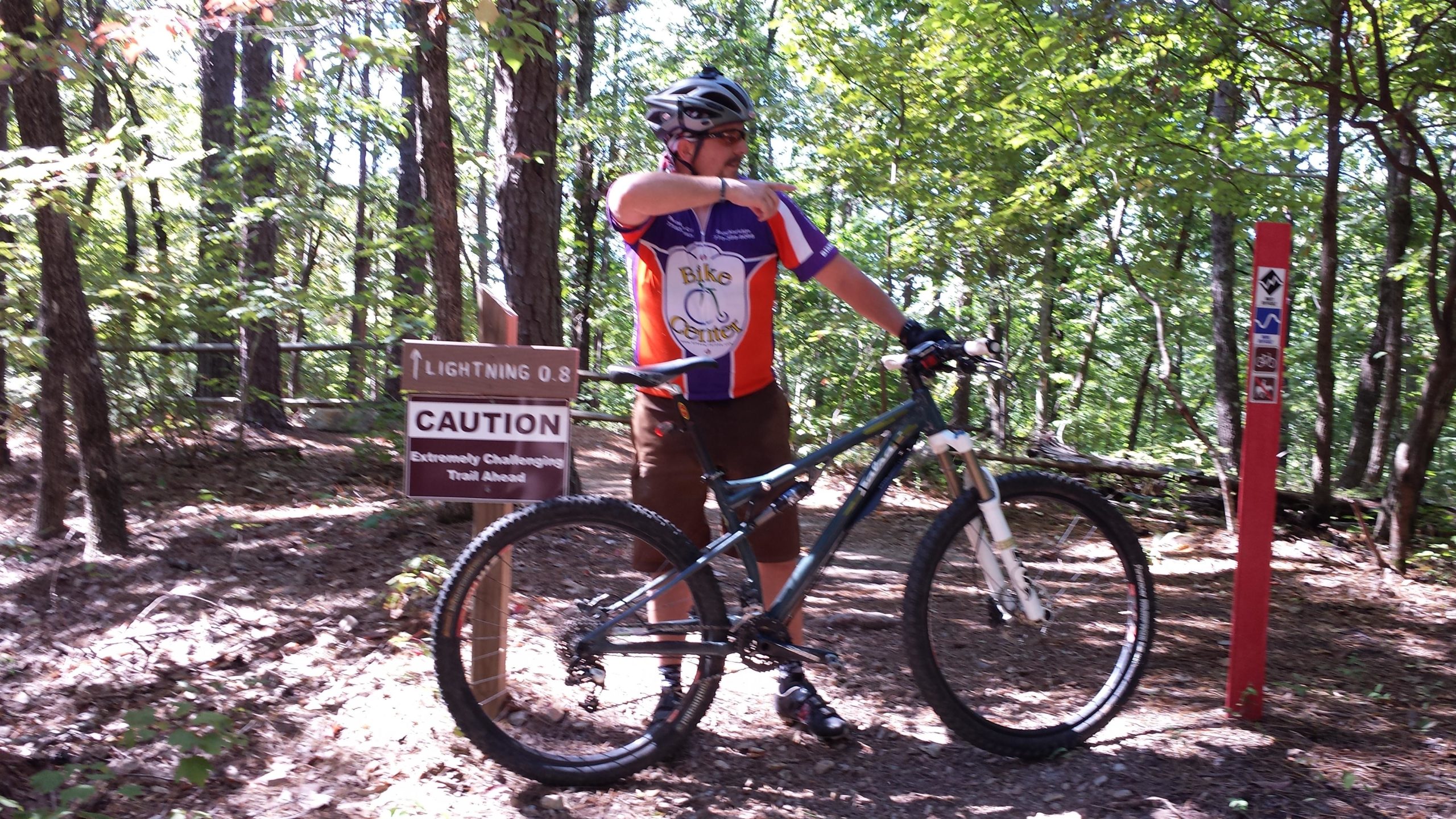 A mountain biker in a colorful jersey stands next to his bike, pointing towards a trail sign that reads "Caution: Extremely Challenging Trail Ahead" with an arrow indicating the direction. The scene is set in a wooded area with trees and foliage in the background. Oak Mountain State Park Bump Trail mountain bike trail.