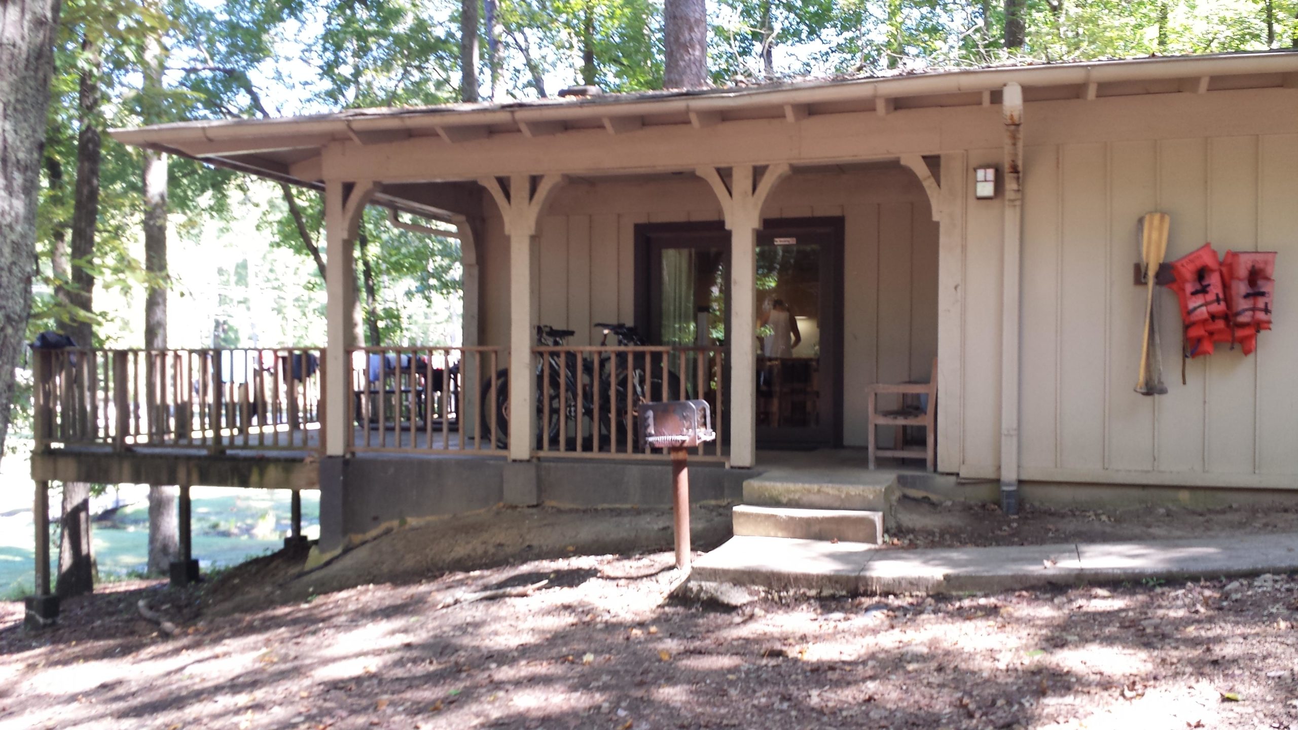 A wooden cabin with a porch surrounded by trees, featuring bicycles leaned against the wall, a small grill on a post, and life jackets hanging on the side. The scene conveys a peaceful outdoor setting. Oak Mountain State Park mountain bike trail.