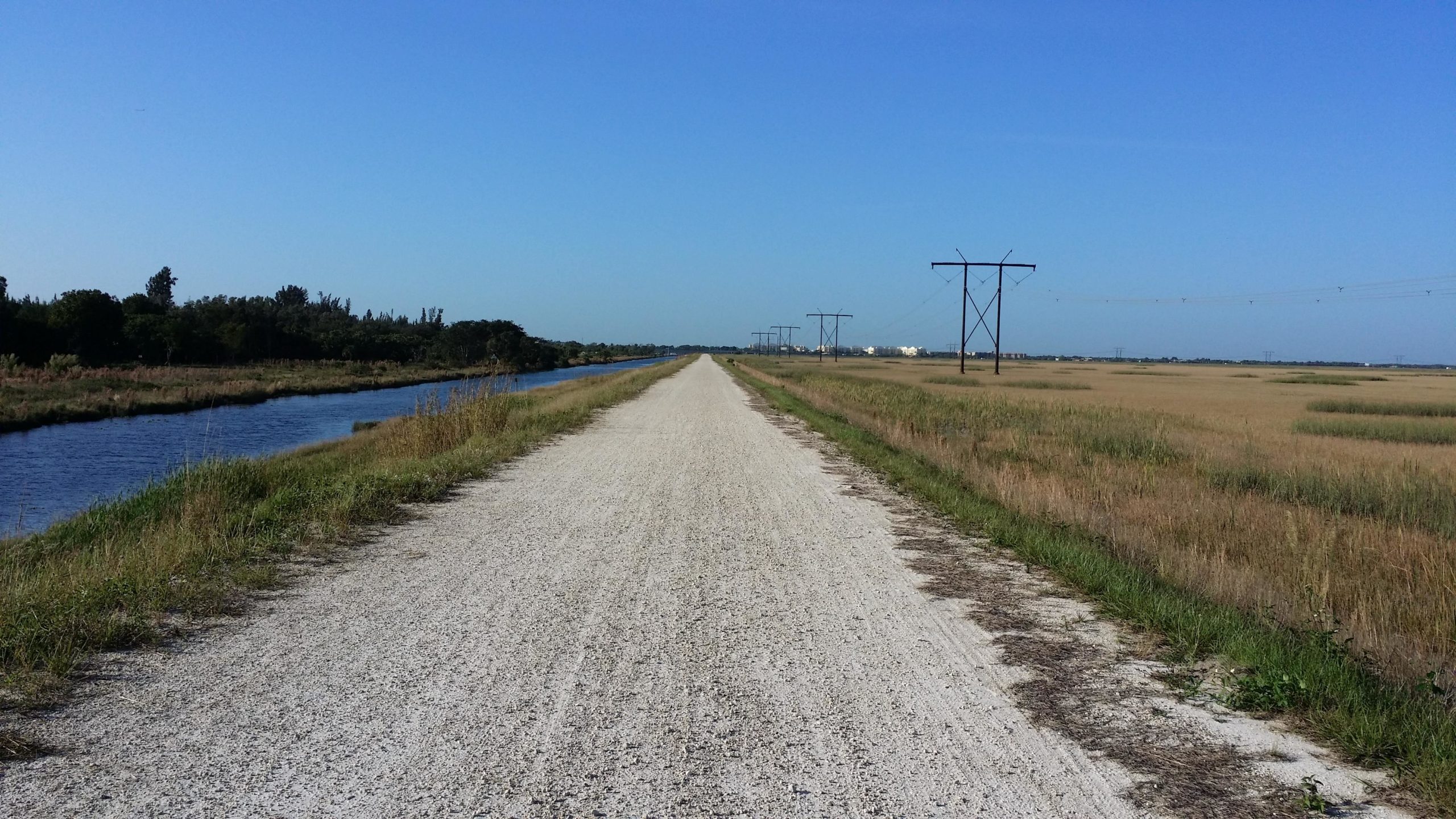 A gravel pathway stretches along a freshwater canal, flanked by tall grasses and trees on one side, with power lines running parallel on the other. In the distance, blue skies are visible above a rural landscape, creating a serene and expansive view. The Levee mountain bike trail.