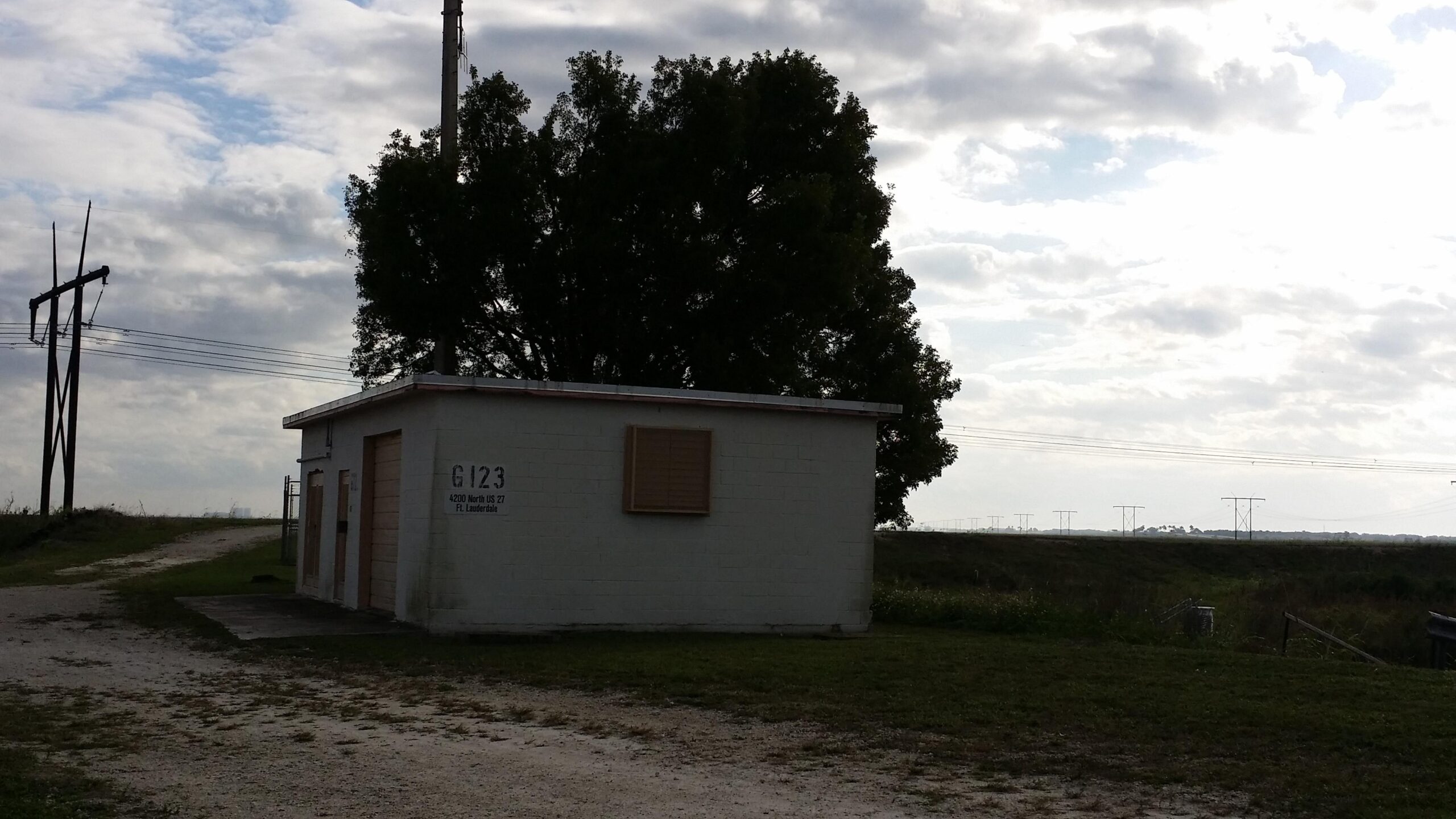 Small building with boarded windows, surrounded by grass and a dirt path, under a cloudy sky with power lines in the background. The Levee mountain bike trail.