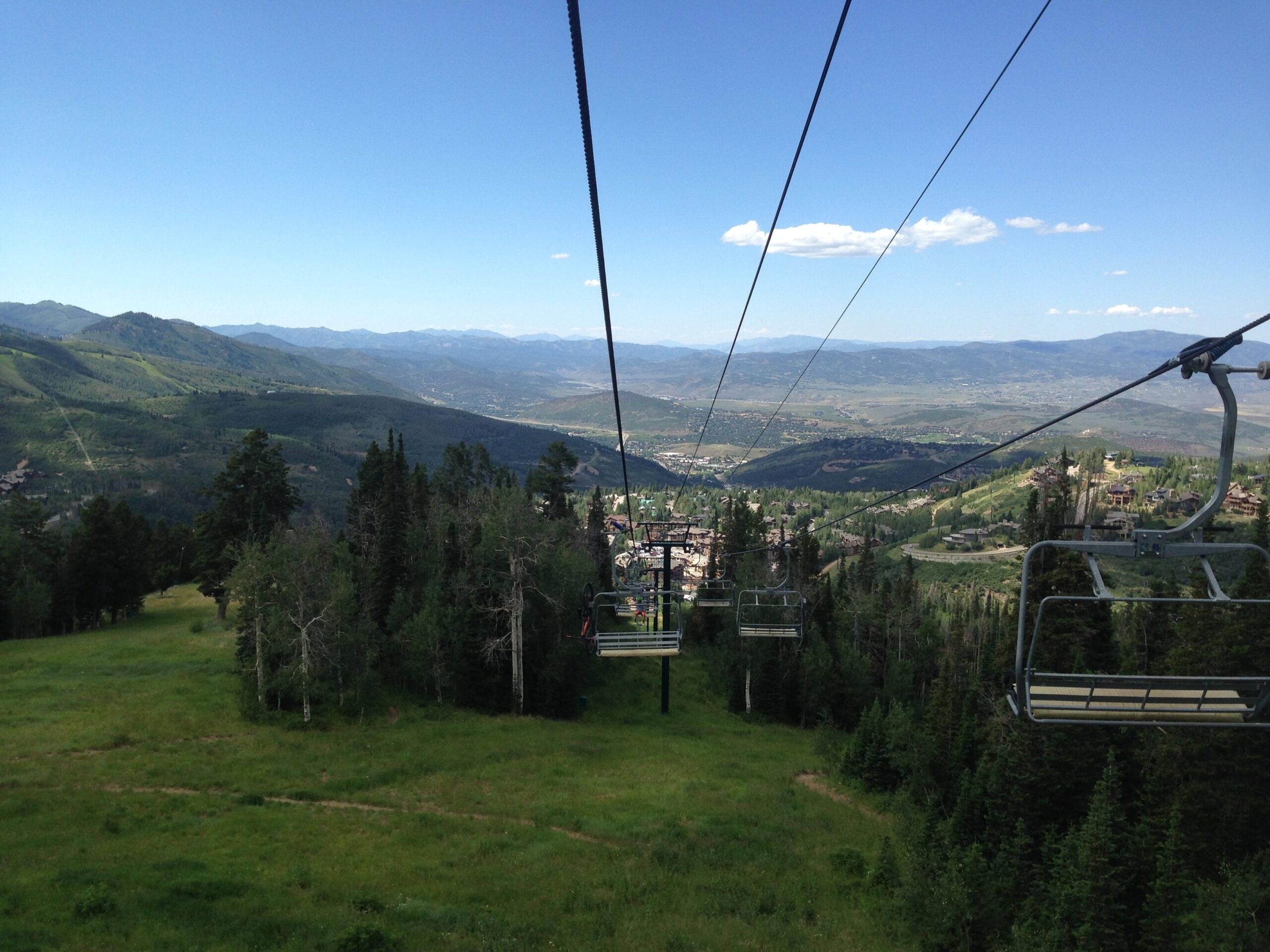 A scenic view from a ski lift, showcasing lush green hills and distant mountains under a clear blue sky. The image captures the chairlift cables and empty seats, with a valley and towns visible in the background, highlighting the beauty of a mountainous landscape. Deer Valley Resort Bike Park mountain bike trail.
