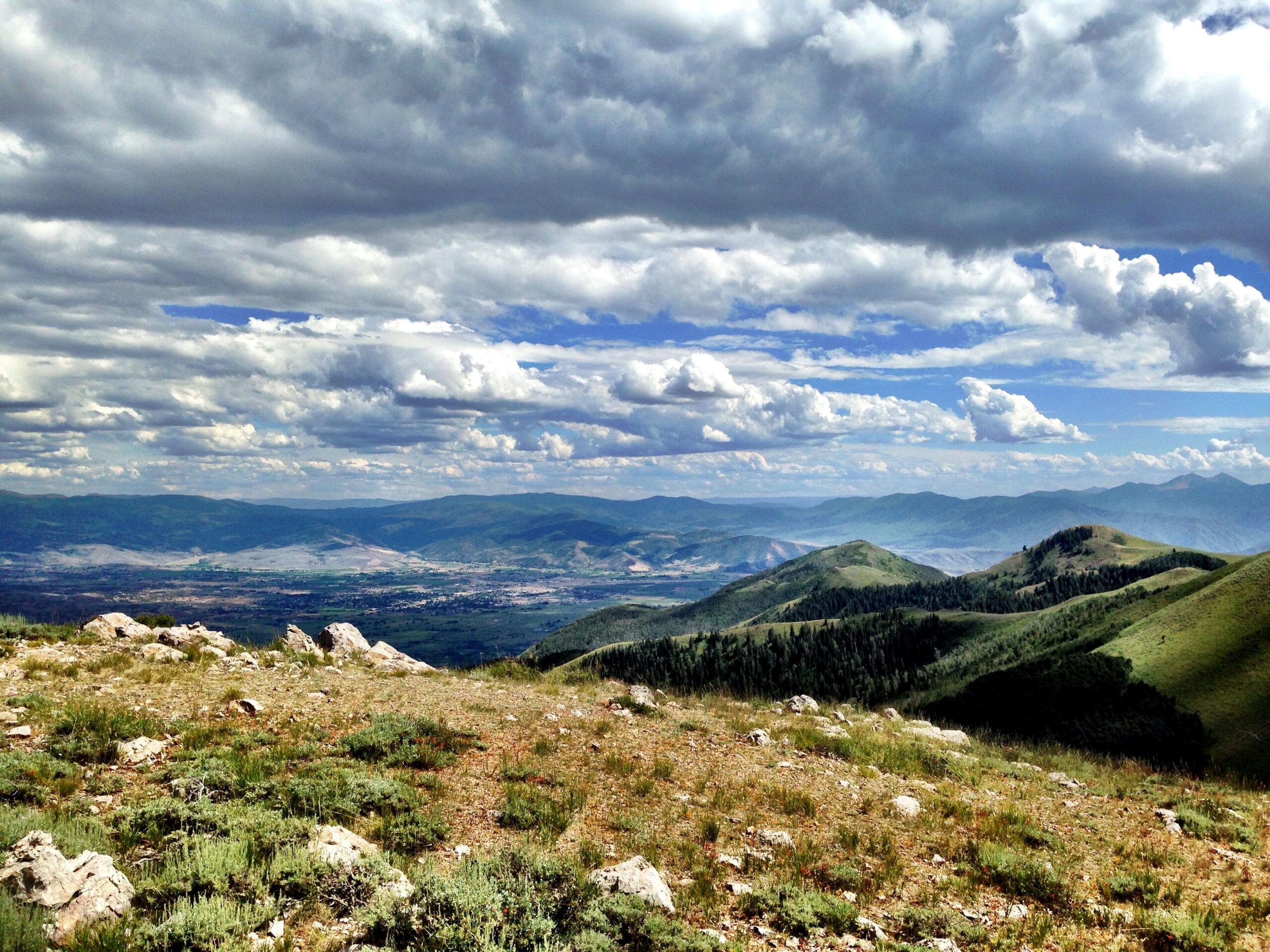 A panoramic view of a mountainous landscape under a partly cloudy sky. Rolling hills are covered with green vegetation and rocky outcrops, while valleys and distant mountains stretch out into the horizon, creating a serene and expansive natural scene. Deer Valley Resort Bike Park mountain bike trail.