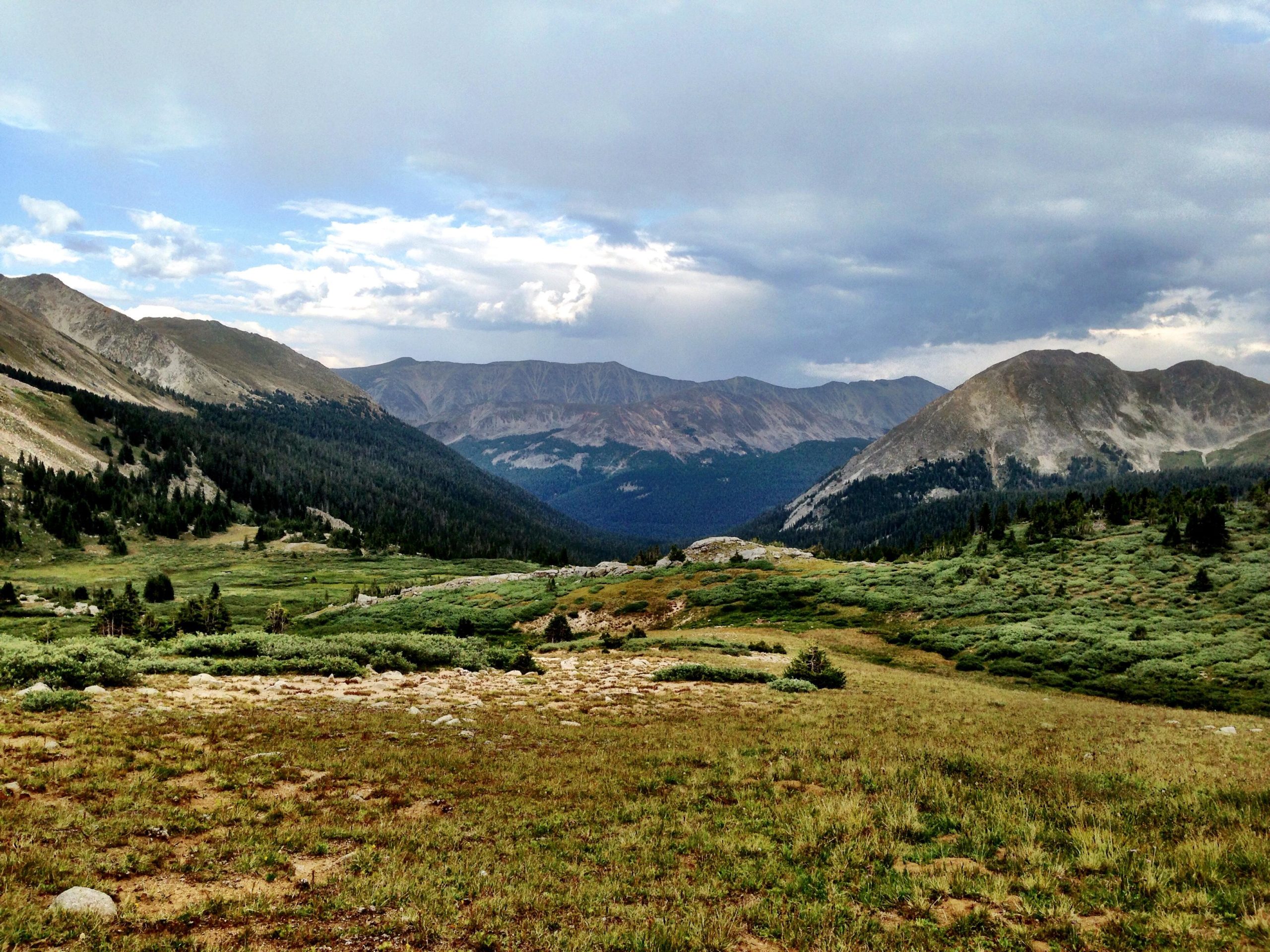 A panoramic view of a mountainous landscape featuring rolling hills, green meadows, and scattered rocky outcrops, under partly cloudy skies. The scene captures the natural beauty and varied terrain of the area, with distant mountains in soft focus. CDT: Alpine Tunnel mountain bike trail.