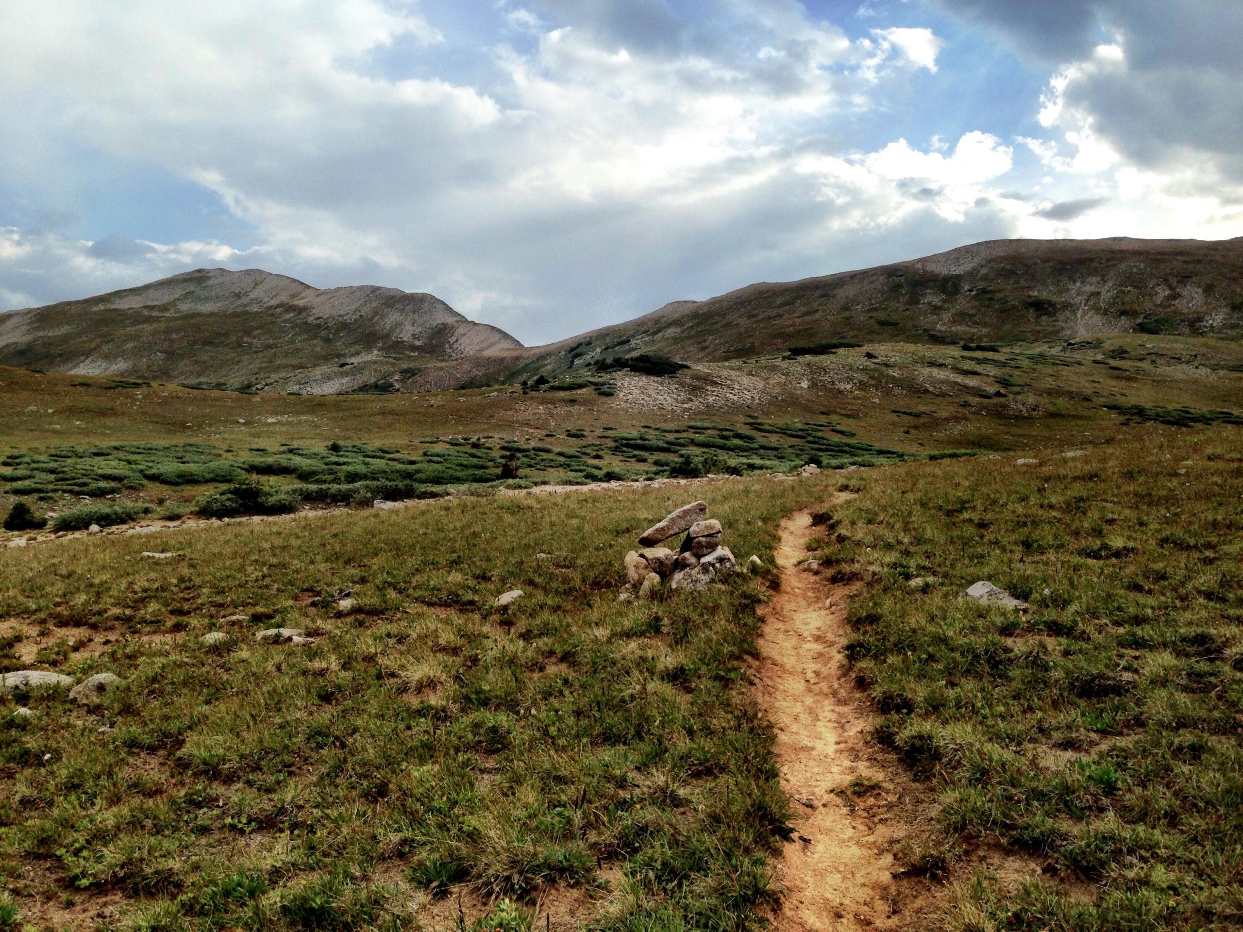 A scenic view of a grassy landscape with a winding dirt path leading through it. In the foreground, there is a small pile of rocks, while rolling hills and mountains rise in the background under a partly cloudy sky. CDT: Alpine Tunnel mountain bike trail.