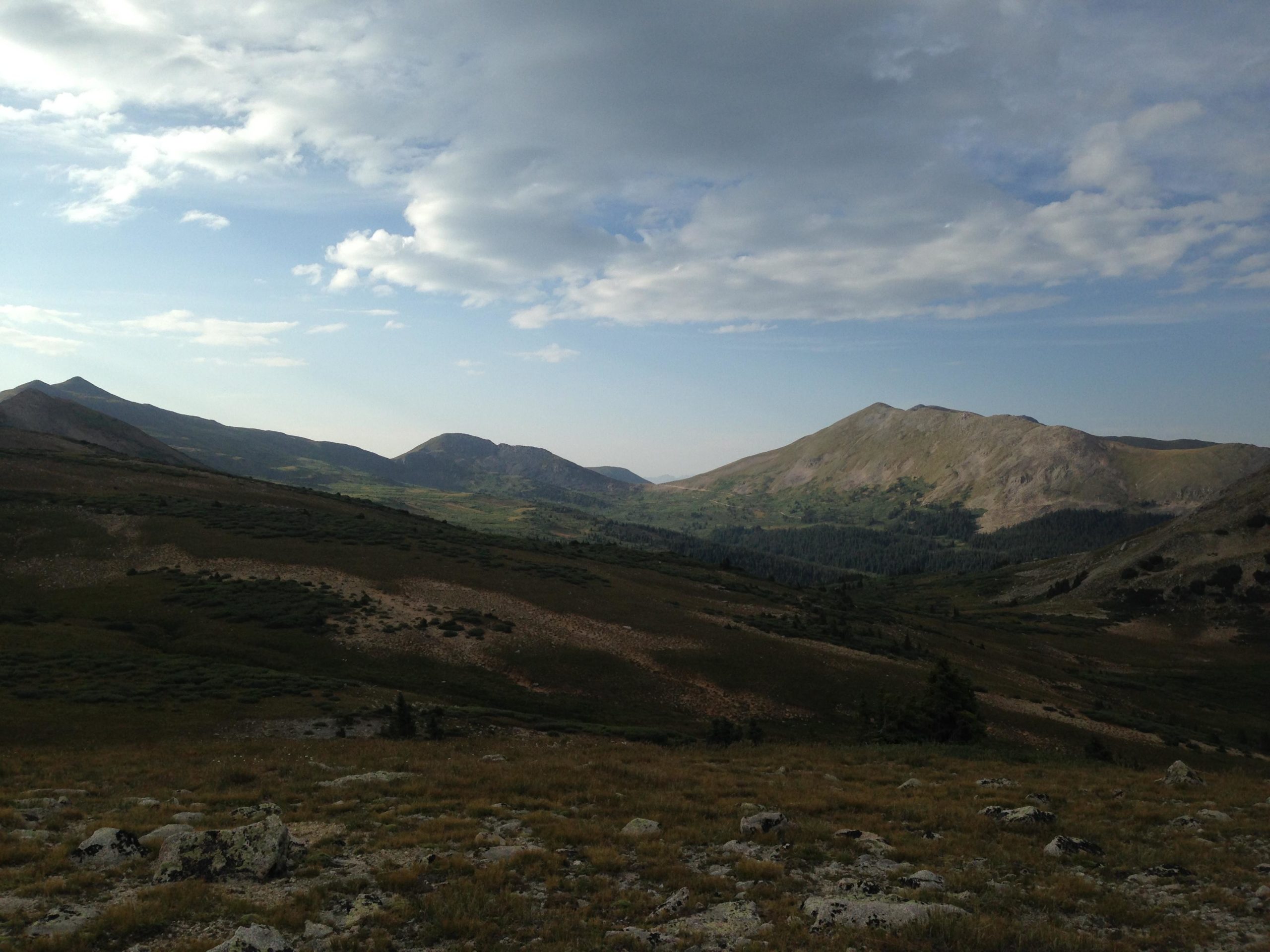 Mountain landscape with rolling hills, grassy plains, and distant peaks under a partly cloudy sky. The scene captures the natural beauty of the terrain with a mix of greenery and rocky features. CDT: Alpine Tunnel mountain bike trail.