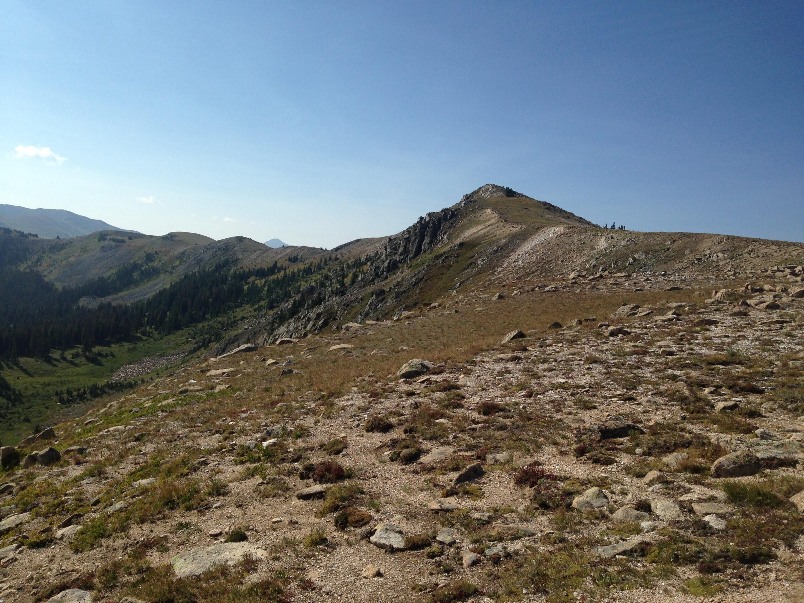 A scenic view of a mountainous landscape under a clear blue sky, featuring rolling hills and rocky outcrops. Green vegetation covers some areas, with a rocky terrain in the foreground. The peaks are partially in shadow, suggesting depth and elevation. Monarch Crest Trail mountain bike trail.