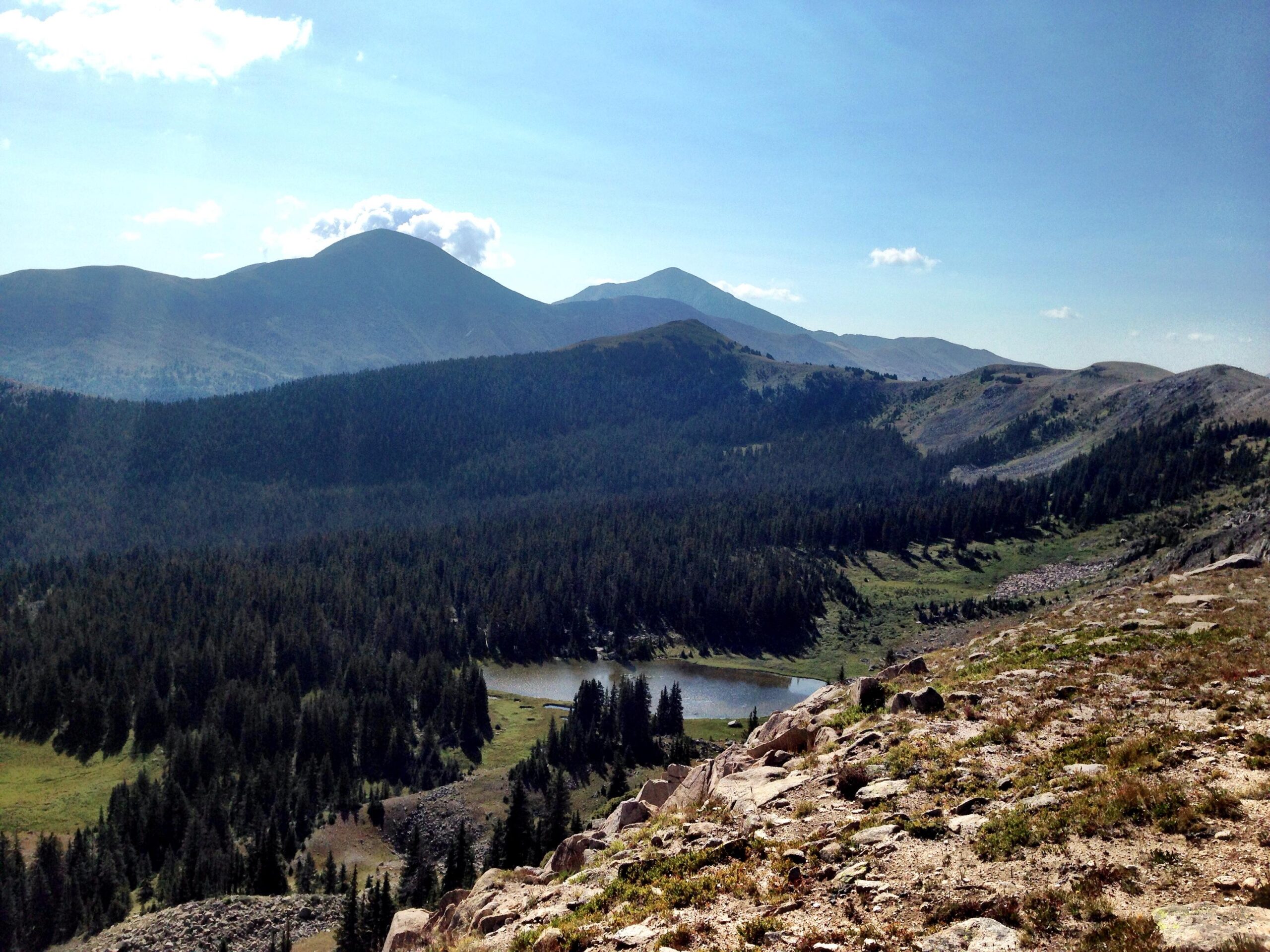 A scenic view of mountainous terrain featuring rolling hills and peaks under a clear blue sky. In the foreground, rocky outcrops and patches of greenery lead to a forested area, while a small lake is visible in the distance, surrounded by trees. The landscape conveys a sense of tranquility and natural beauty. Monarch Crest Trail mountain bike trail.