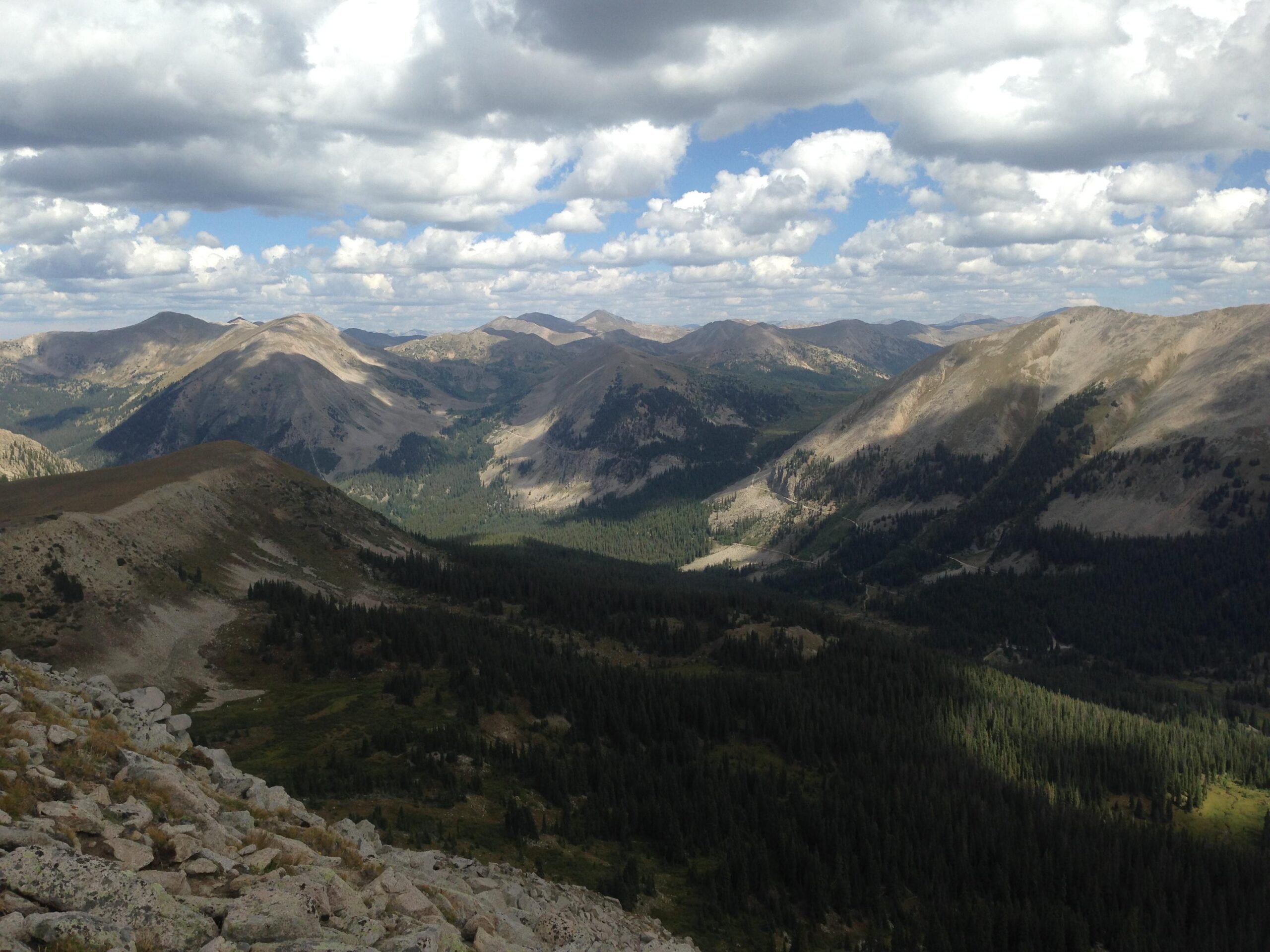 A scenic view of mountains under a partly cloudy sky. The landscape features rolling hills and valleys, a mix of rocky slopes and lush green forests, capturing the beauty of nature in a mountainous region. Canyon Creek Trail mountain bike trail.