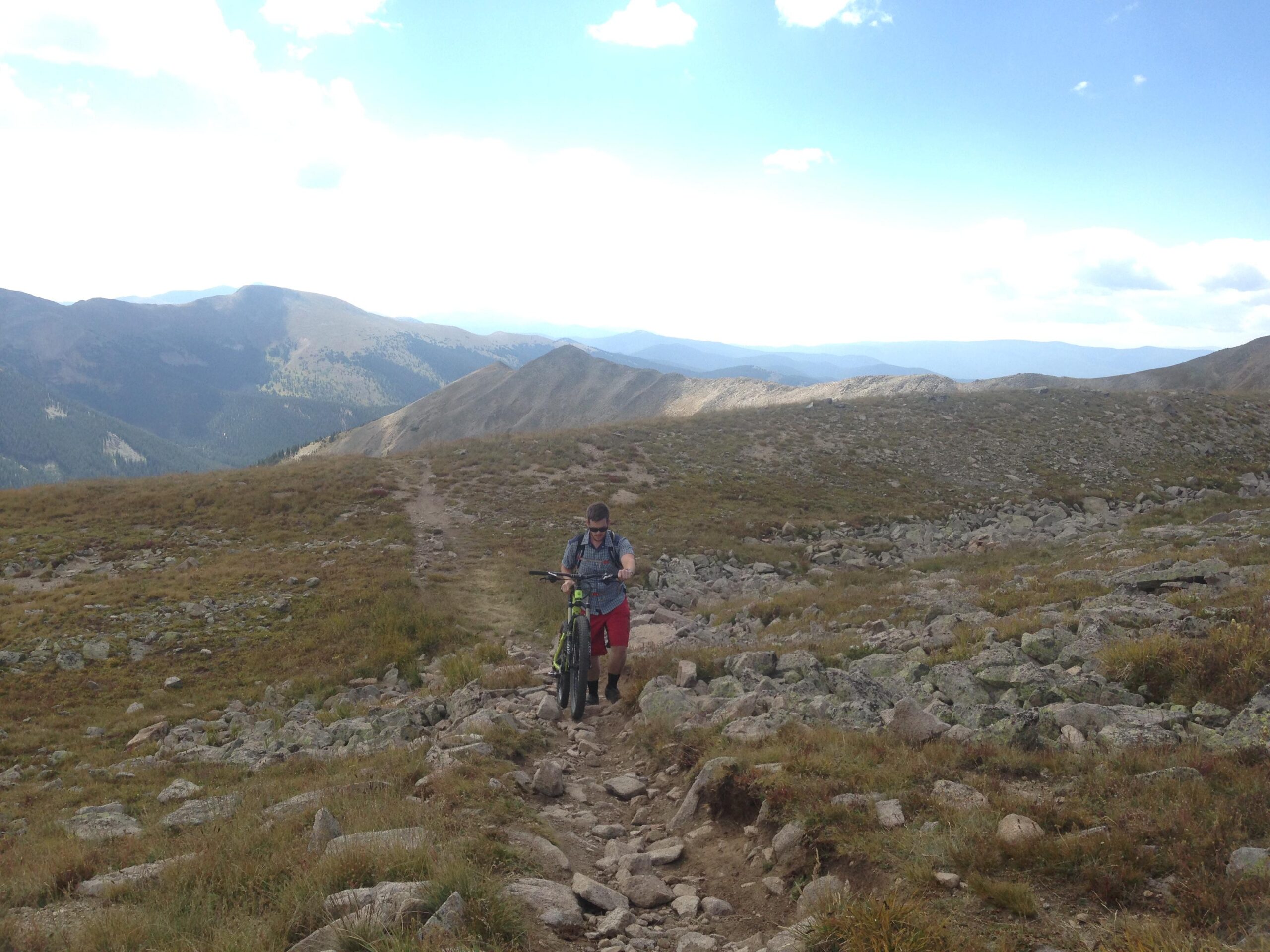 A mountain biker walking with their bike along a rocky trail in a mountainous area, surrounded by grassy hills and distant peaks under a partly cloudy sky. Canyon Creek Trail mountain bike trail.