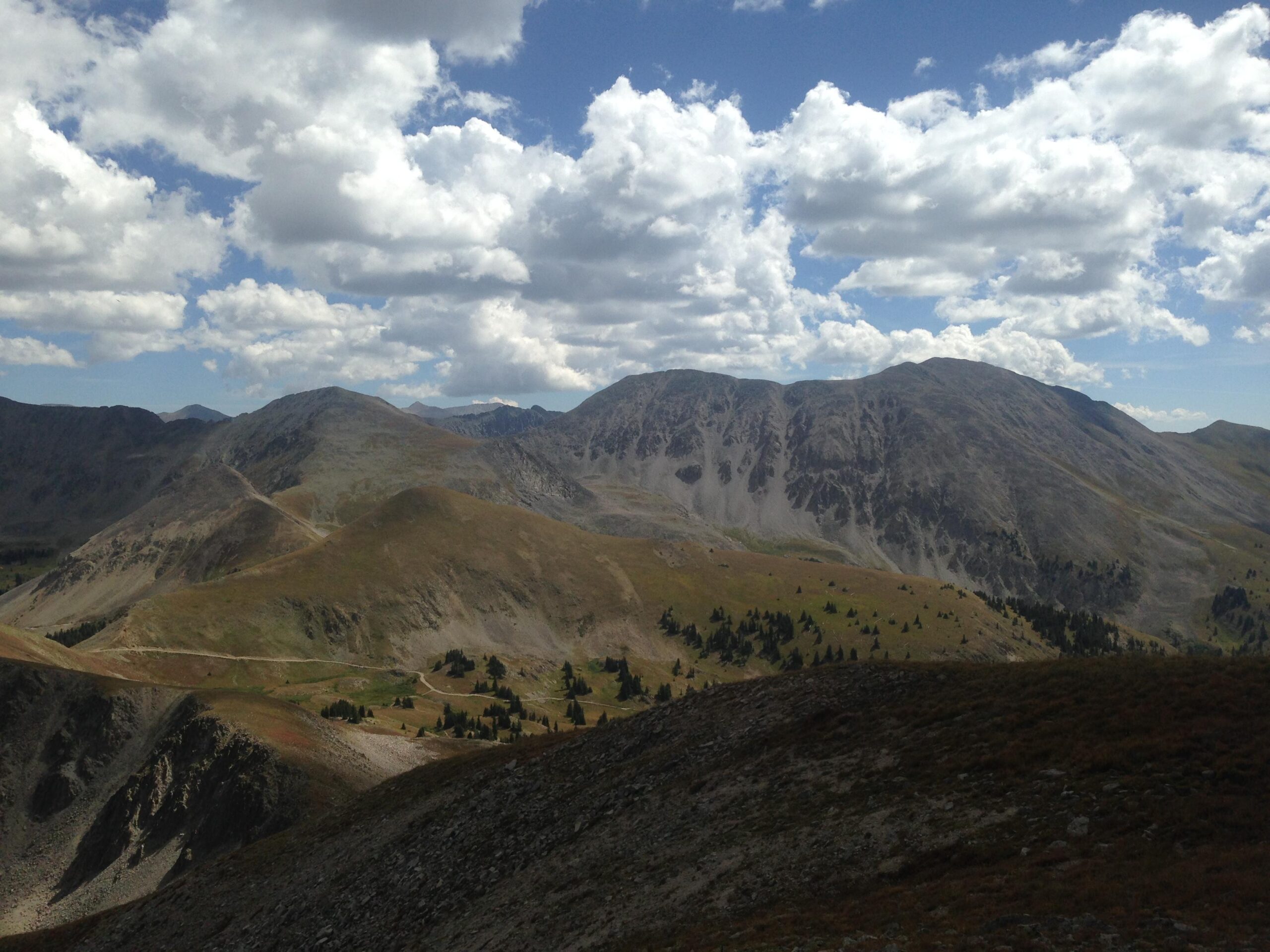 A panoramic view of rugged mountains under a mostly cloudy sky, featuring rolling hills and patches of greenery in the foreground with rocky slopes and peaks in the background. Canyon Creek Trail mountain bike trail.