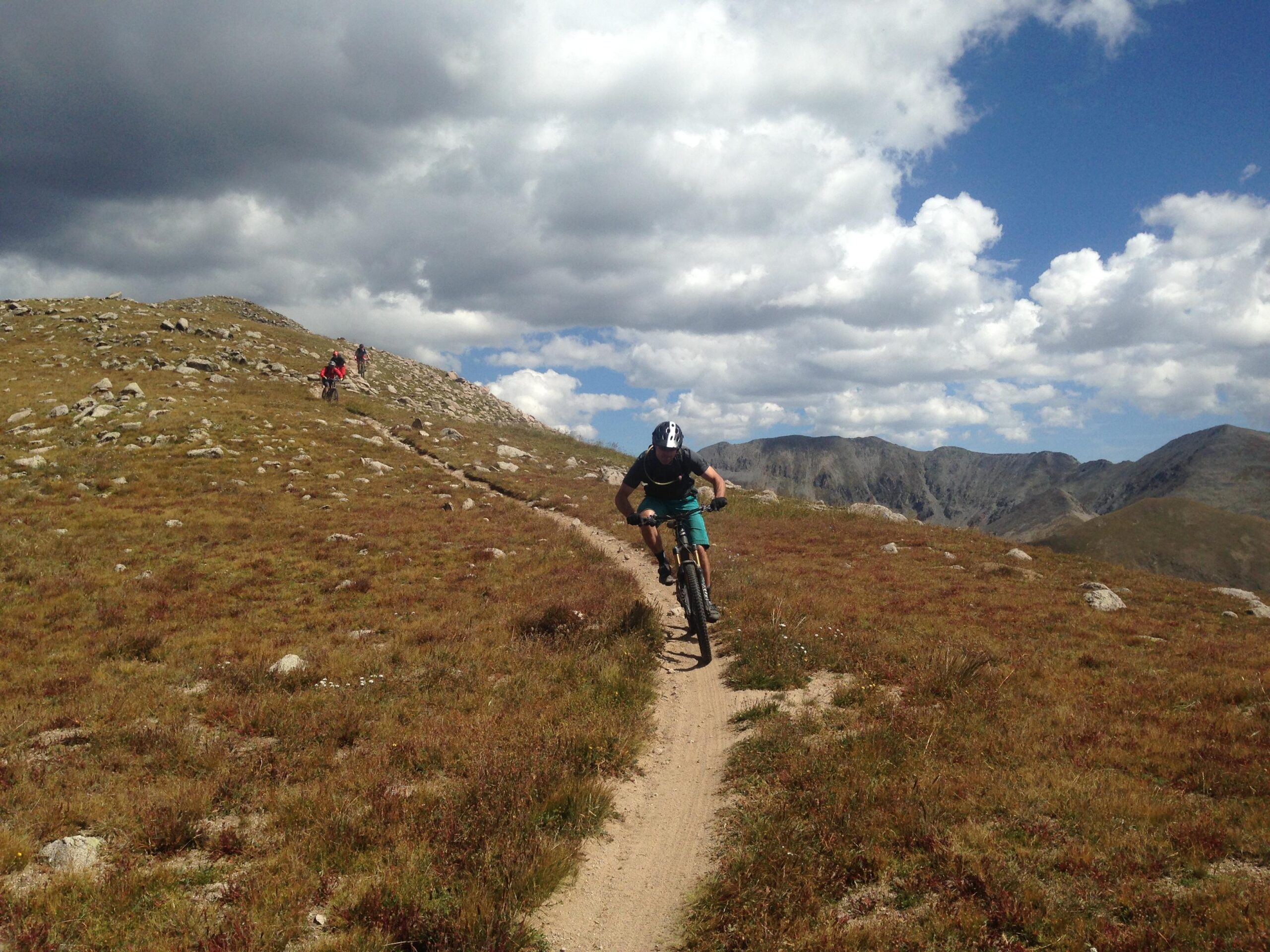 A mountain biker rides along a narrow dirt trail amidst a rugged landscape, with grassy hills and rocks visible in the foreground. In the background, mountains rise under a partly cloudy sky. Another cyclist can be seen in the distance. Canyon Creek Trail mountain bike trail.