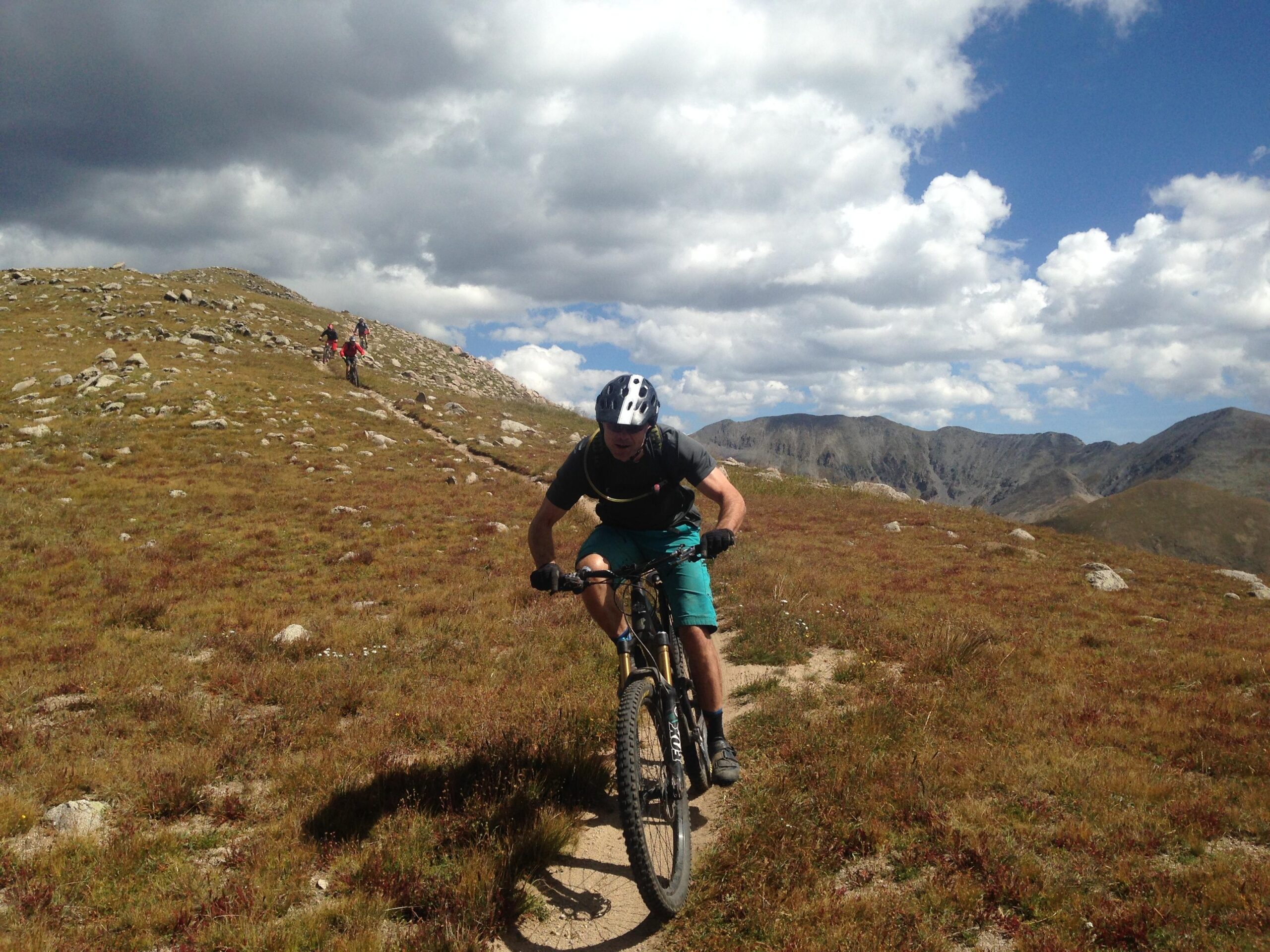 A mountain biker navigating a trail through a rocky landscape, with grass and scattered stones. In the background, other cyclists are seen climbing a hill under a cloudy sky with a mountainous horizon. Canyon Creek Trail mountain bike trail.