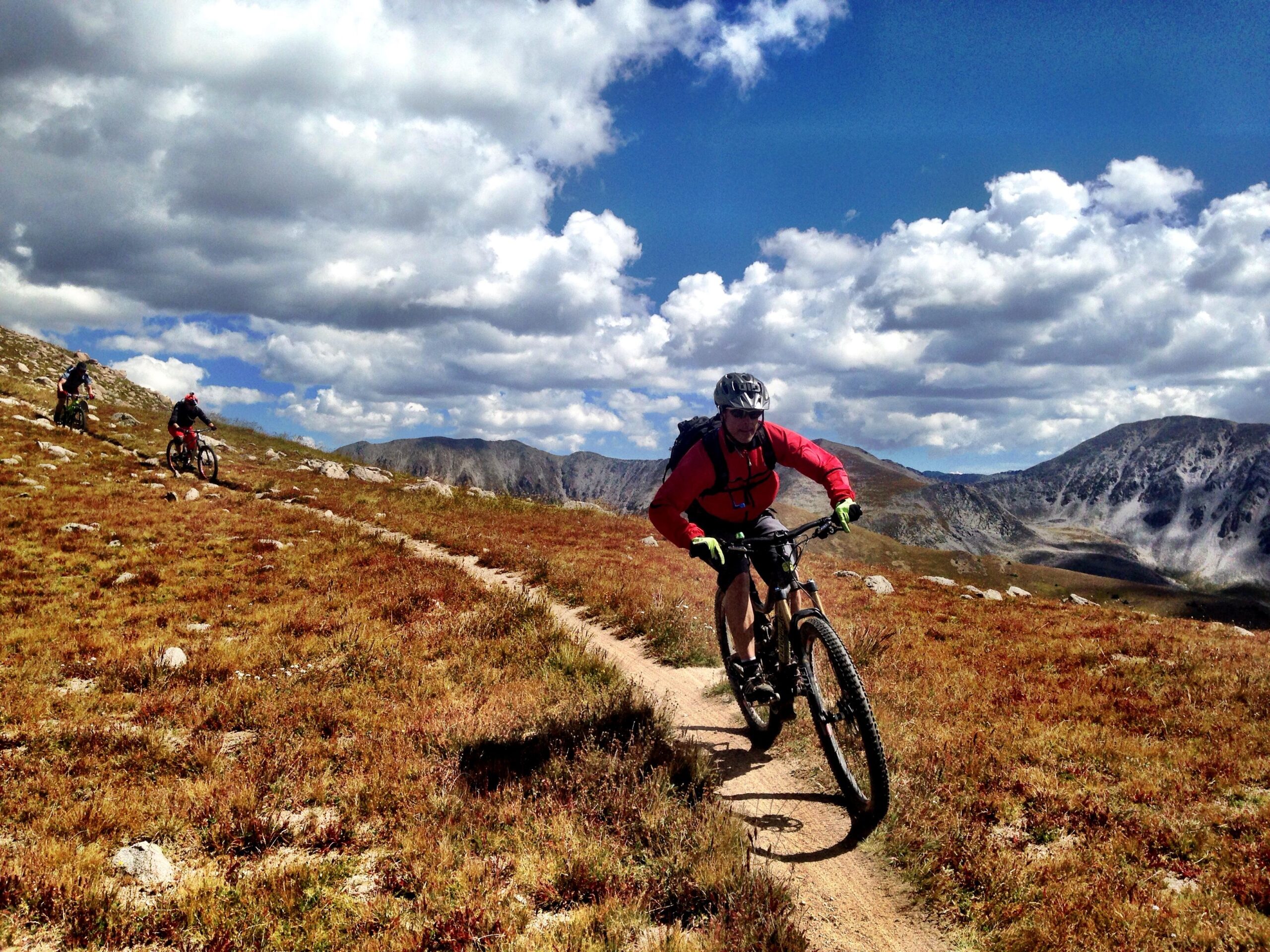 A group of mountain bikers rides along a narrow dirt trail in a grassy landscape with rocky patches, surrounded by mountains and a partly cloudy blue sky. One biker in a red jacket is prominently featured, focused on navigating the trail. Canyon Creek Trail mountain bike trail.