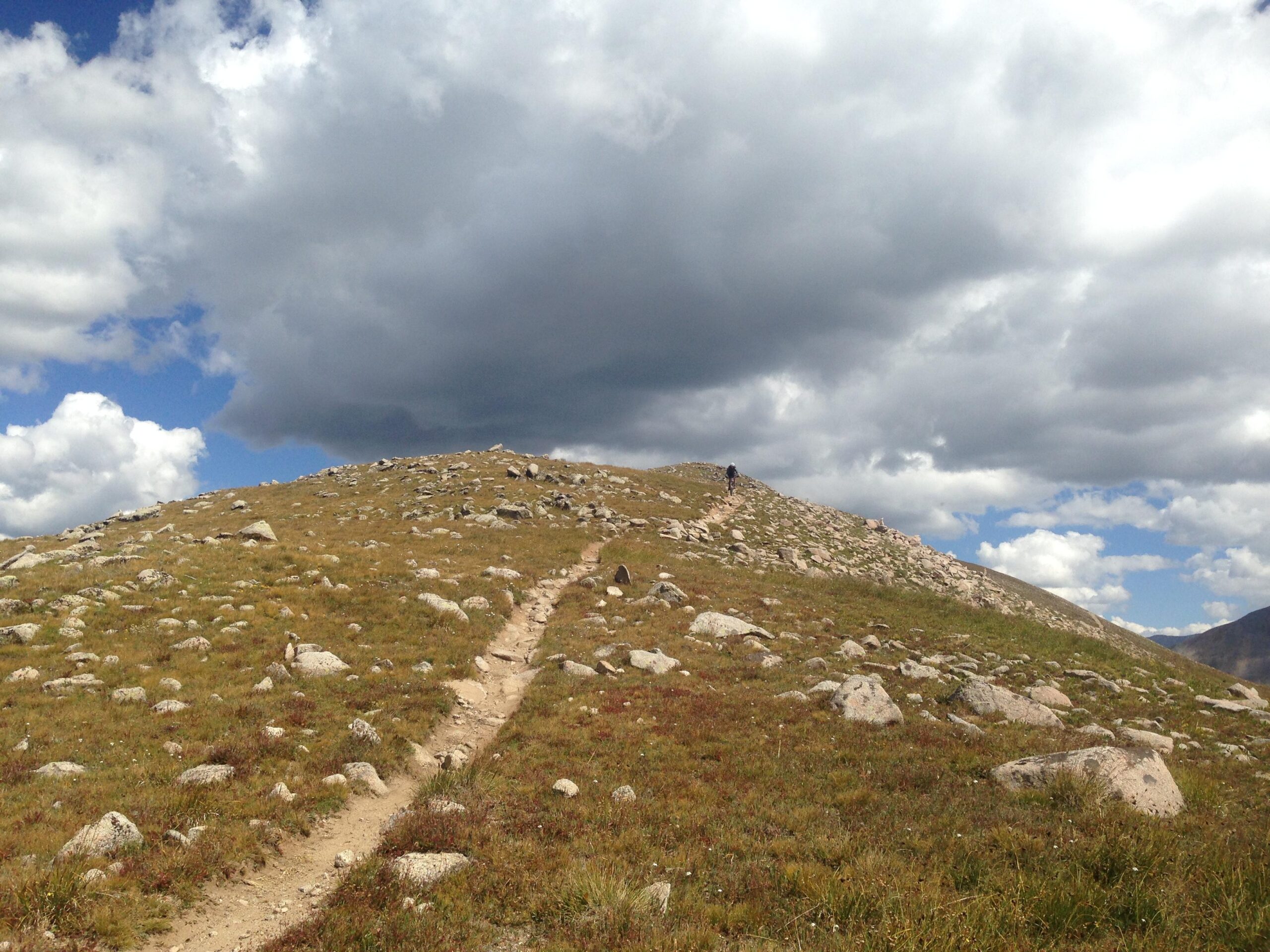 A hiker walks along a winding dirt path on a grassy hill covered with rocks, beneath a partly cloudy sky. The landscape features a gentle slope leading towards a summit, with patches of blue peeking through the clouds. Canyon Creek Trail mountain bike trail.