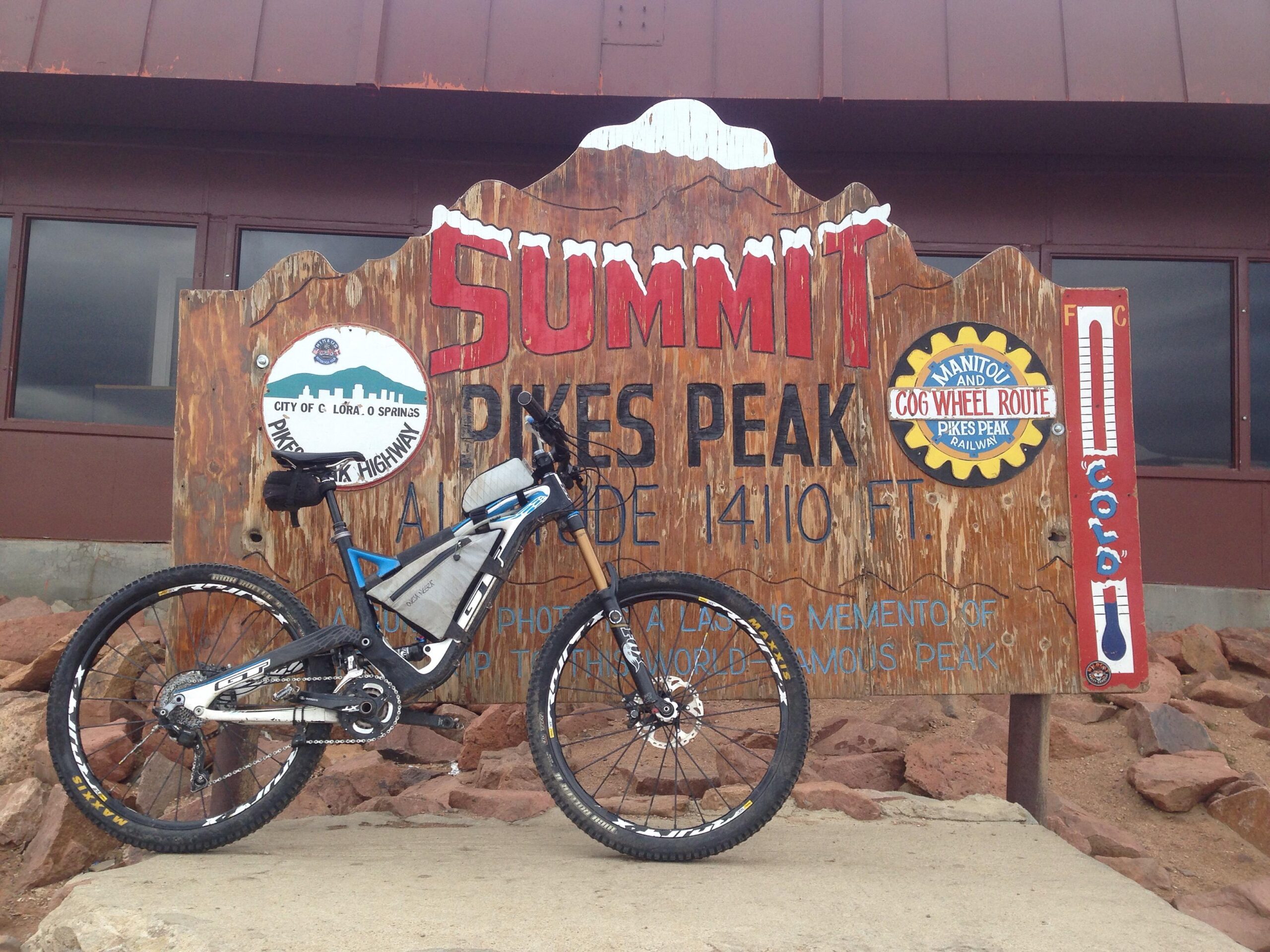 A mountain bike is parked in front of a weathered wooden sign at the summit of Pikes Peak, indicating an elevation of 14,110 feet. The sign features various icons and text, including “Summit,” the city of Colorado Springs, and information about the Pikes Peak Highway and the Manitou and Cog Wheel Route. The background shows a building with large windows. Barr Trail / Pikes Peak mountain bike trail.