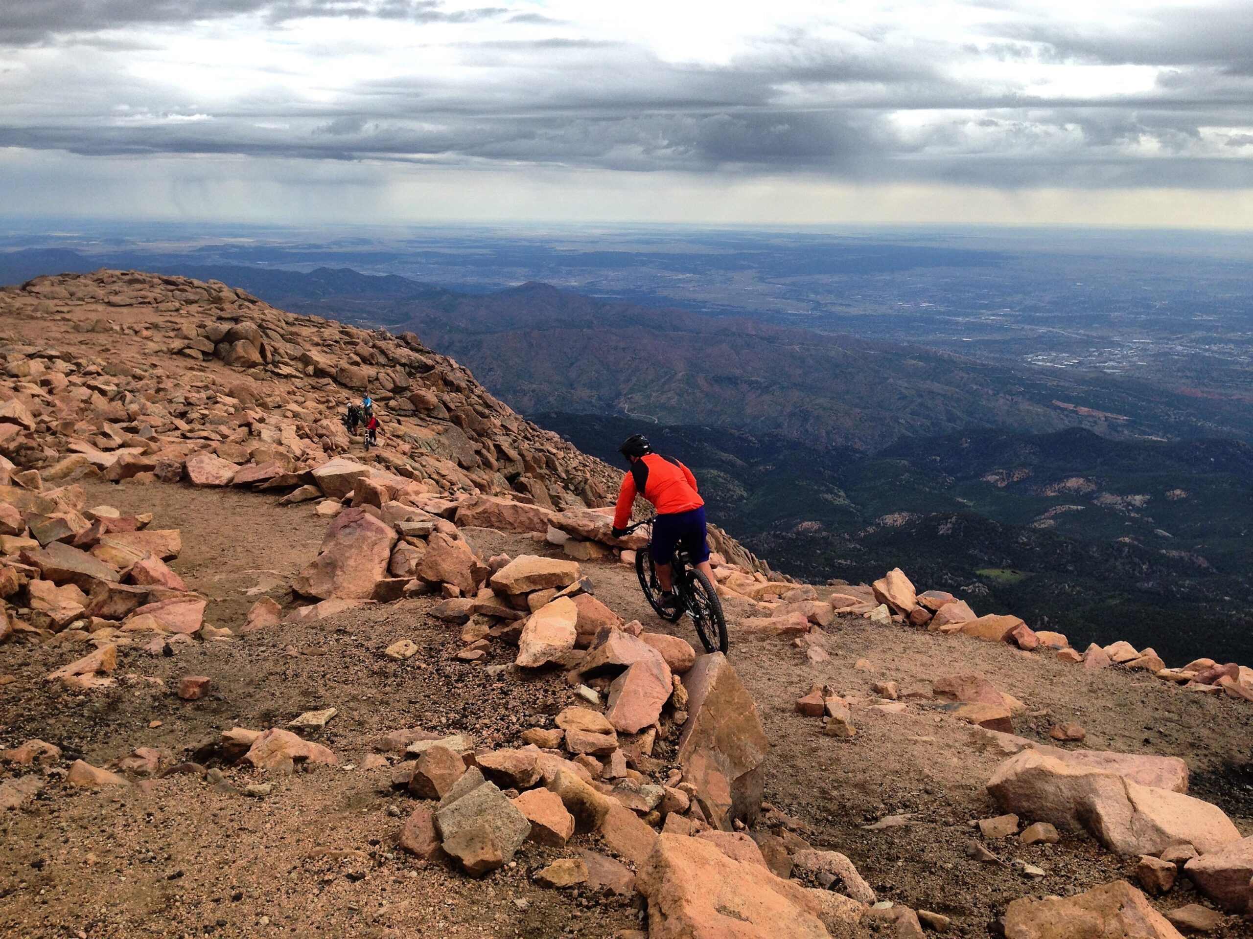 A mountain biker navigates a rocky trail on a mountainous landscape, with layers of hills and valleys visible in the background under an overcast sky. The scene captures the rugged terrain and adventurous spirit of outdoor biking. Barr Trail / Pikes Peak mountain bike trail.