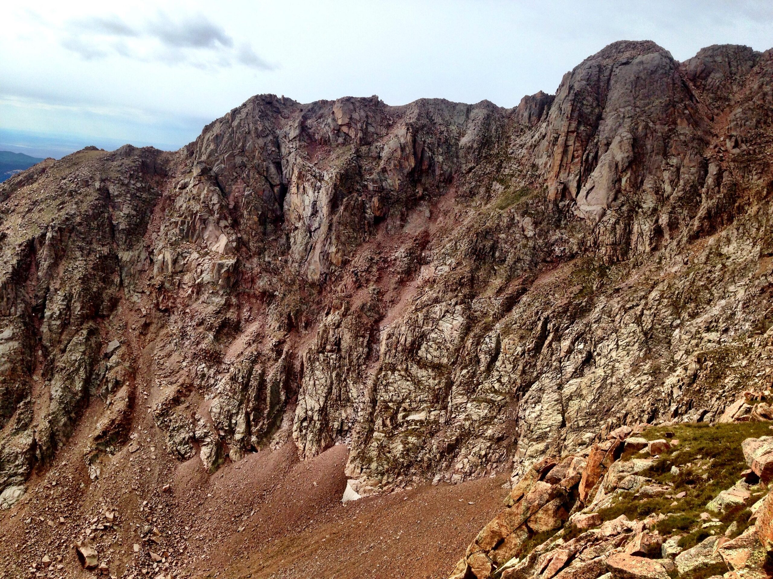 A rugged mountain landscape featuring steep, rocky cliffs and craggy terrain. The scene includes a mix of textured rock faces and scattered boulders, with a cloudy sky above and a sense of wilderness. Barr Trail / Pikes Peak mountain bike trail.