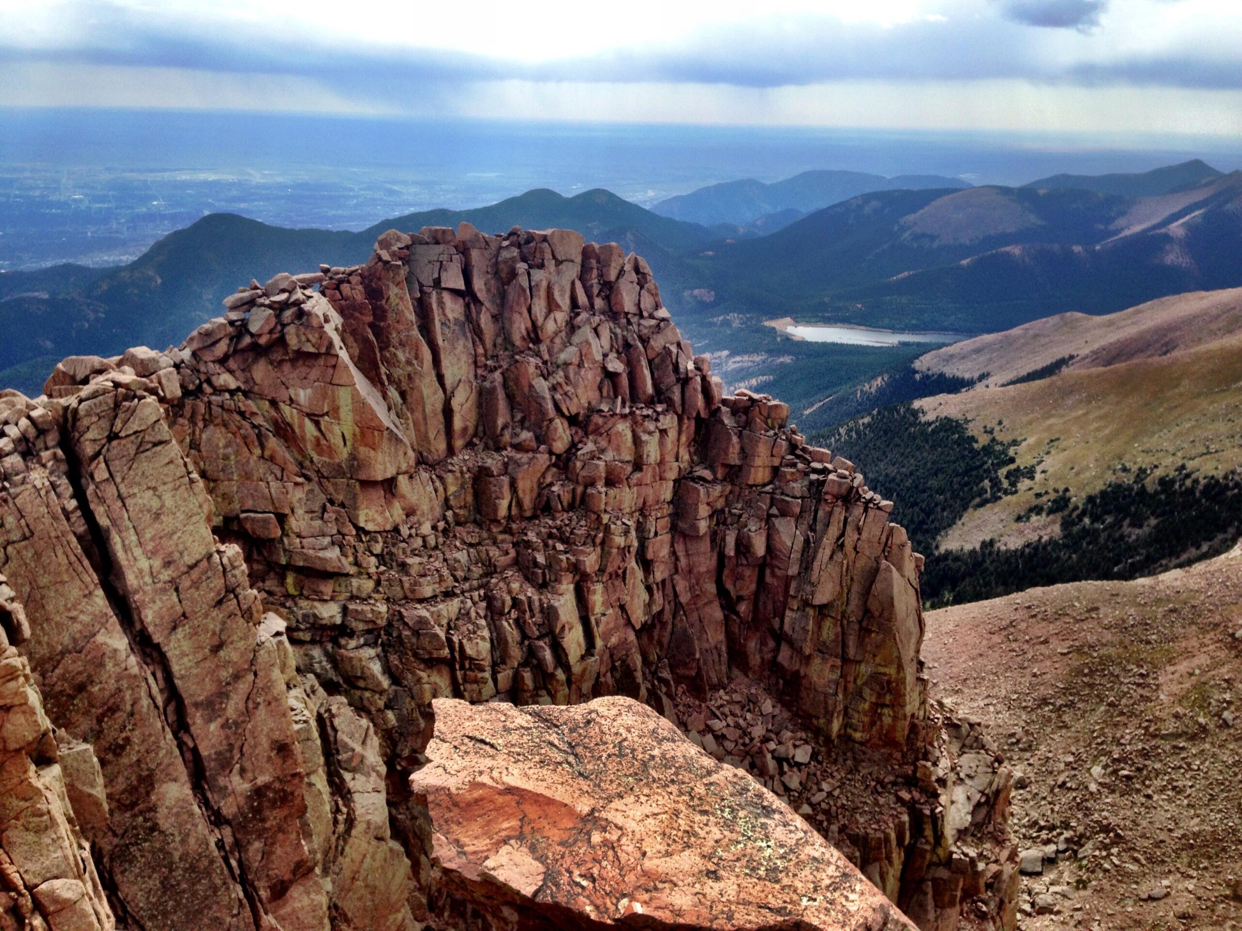 A panoramic view from a rocky mountain summit, featuring rugged cliffs in the foreground and rolling hills in the background. The landscape is dotted with patches of greenery and a body of water can be seen nestled among the hills. The sky is overcast, adding a dramatic feel to the scene. Barr Trail / Pikes Peak mountain bike trail.