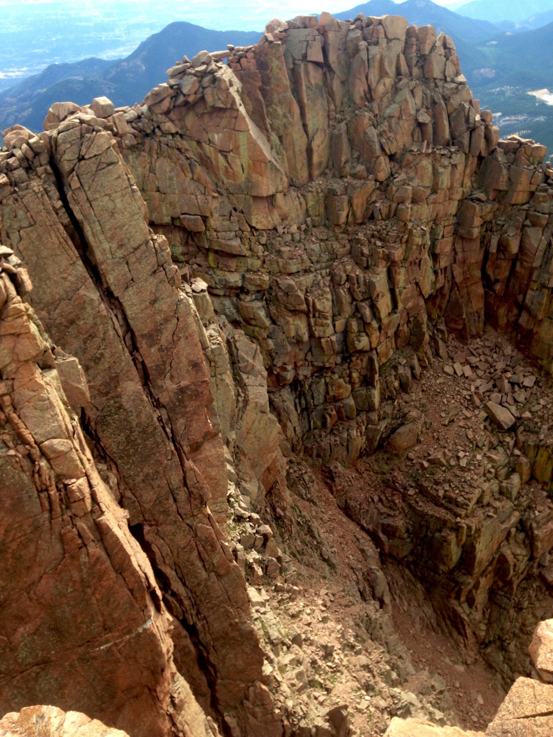 A steep rocky cliff with vertical layers and jagged edges, extending downward into a rocky gorge. The background features distant mountains and a clear blue sky. The scene captures the ruggedness of the landscape. Barr Trail / Pikes Peak mountain bike trail.