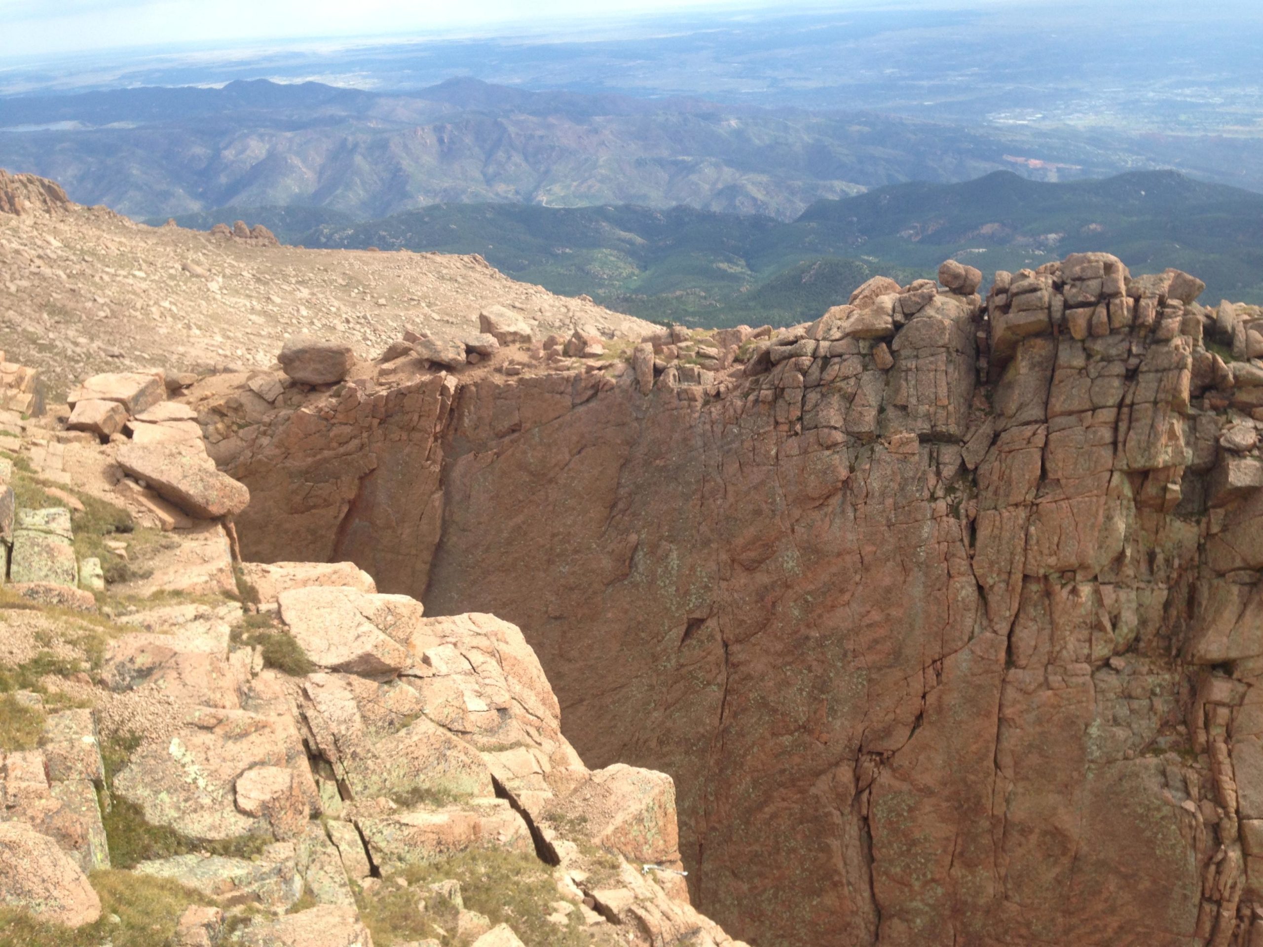 A rocky mountain landscape with steep cliffs and a deep gorge, extending into distant green hills and valleys under a clear sky. The foreground features large, textured granite boulders and sparse vegetation. Barr Trail / Pikes Peak mountain bike trail.