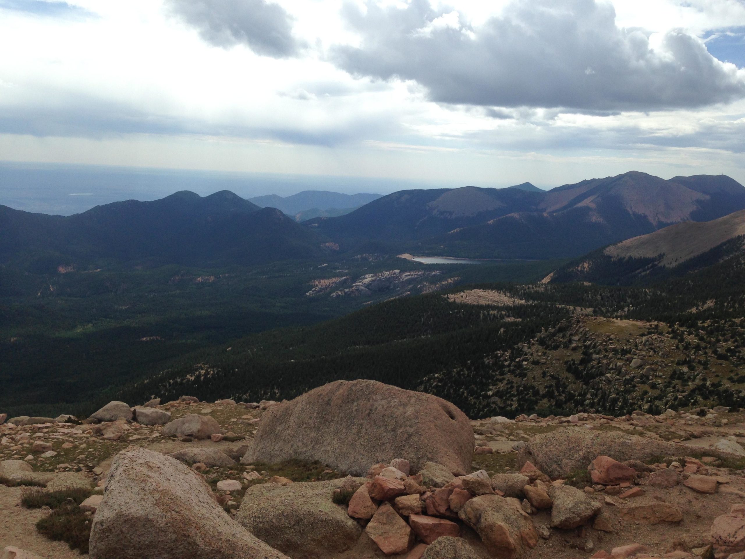 A panoramic view of mountainous terrain under a partly cloudy sky. The landscape features rolling green hills, rocky outcrops, and distant peaks, with patches of forest and a small body of water visible in the valley below. Barr Trail / Pikes Peak mountain bike trail.