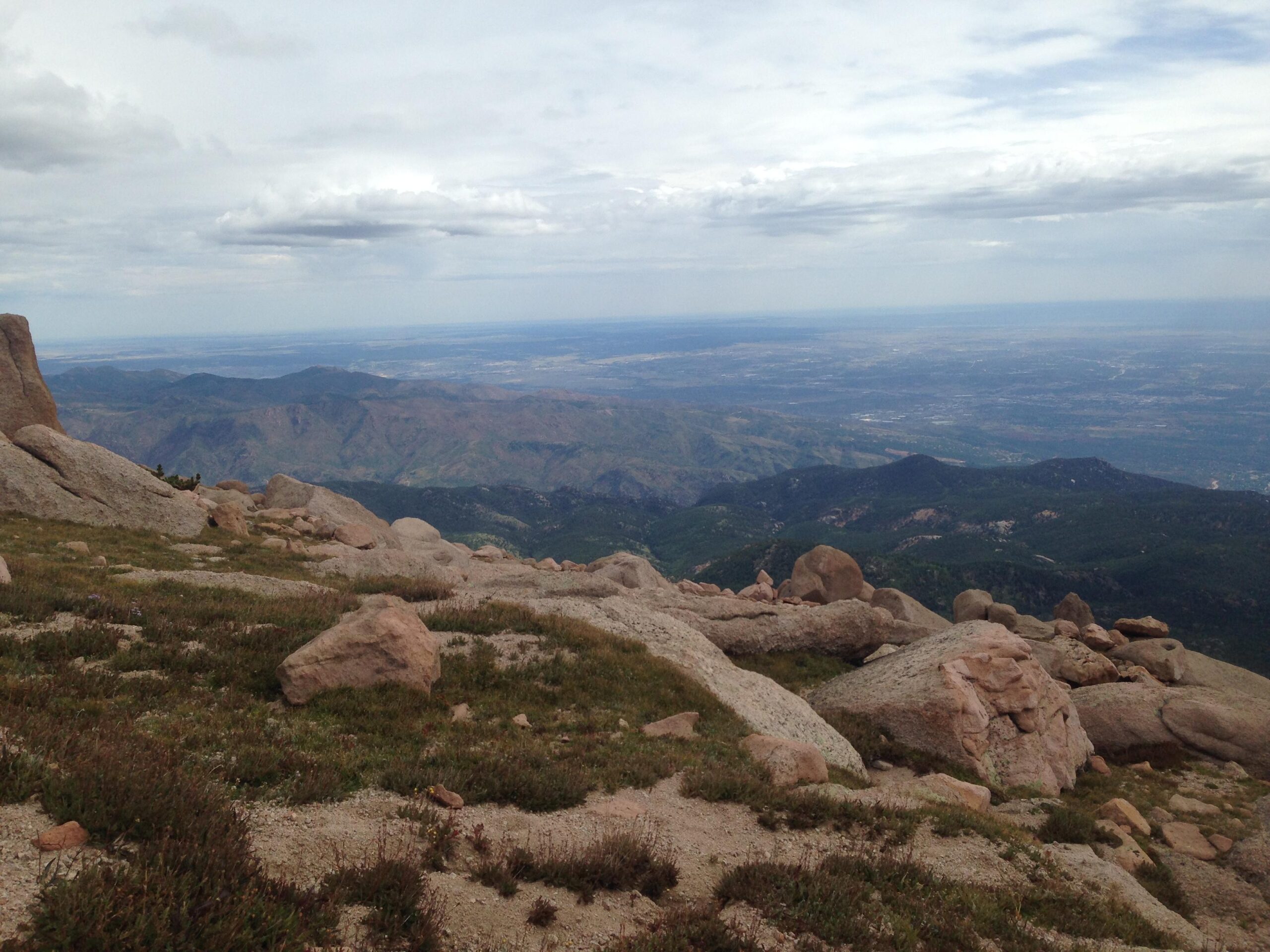 A panoramic view from a rocky mountain peak, showcasing a vast landscape of rolling hills and valleys below. The foreground features large boulders and patches of grass, while the distant mountains fade into the horizon under a cloudy sky. Barr Trail / Pikes Peak mountain bike trail.