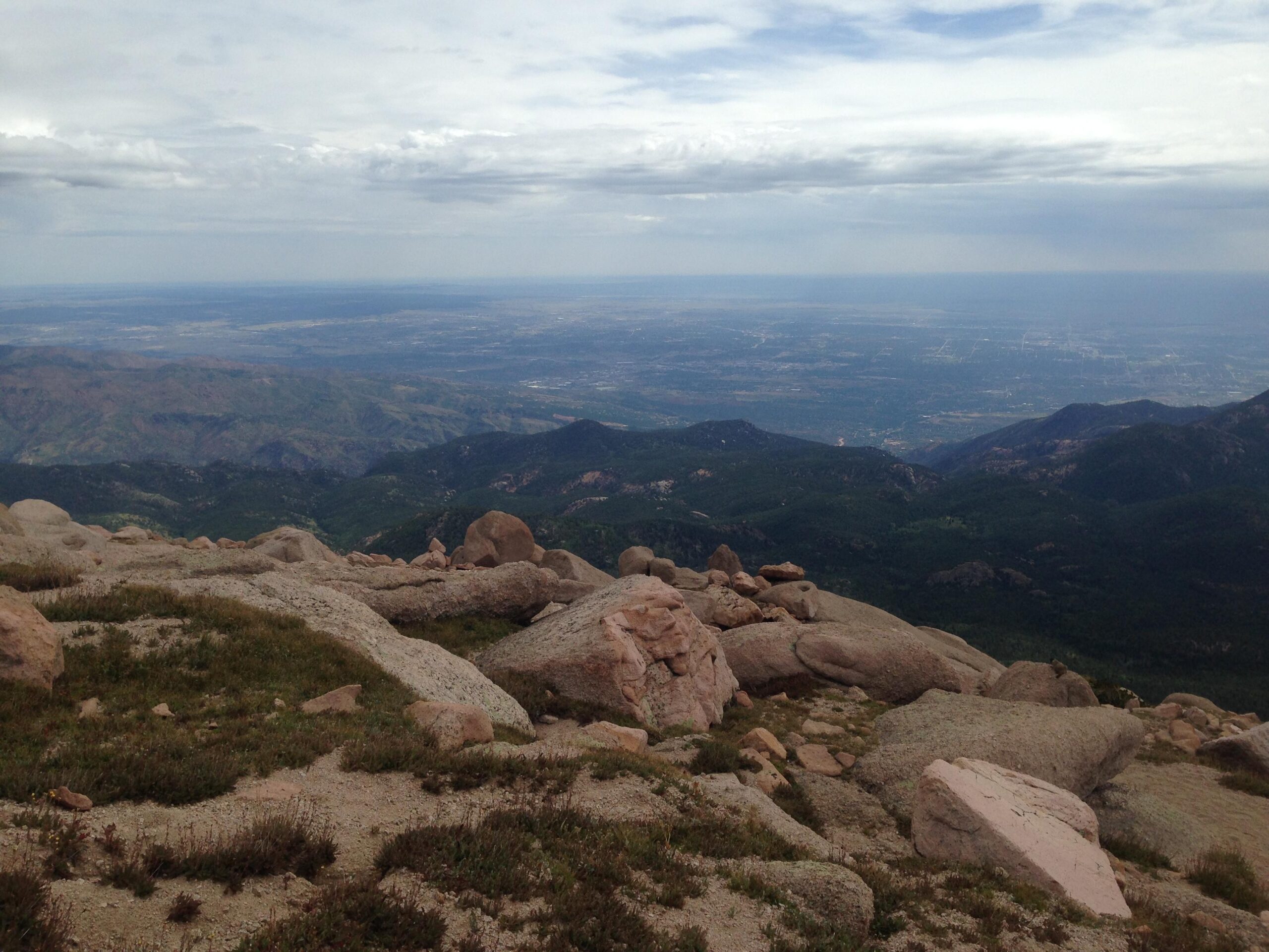 A scenic view from a mountaintop, showcasing rocky terrain and lush green hills stretching into the distance. The sky is partly cloudy, with expansive views of the landscape below, including valleys and distant mountains. Barr Trail / Pikes Peak mountain bike trail.