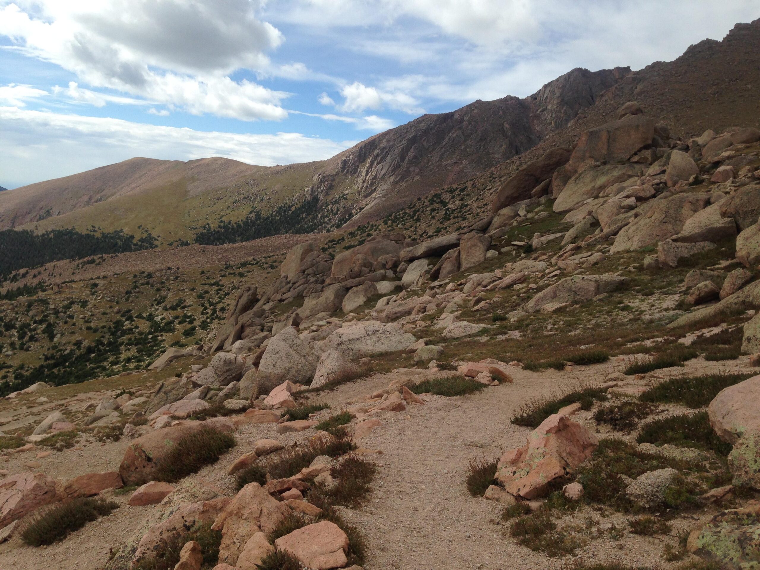 A rocky hillside with a winding trail, surrounded by grassy patches and sparse vegetation, under a partly cloudy sky. The landscape features rolling mountains in the background, providing a scenic view of nature's rugged terrain. Barr Trail / Pikes Peak mountain bike trail.