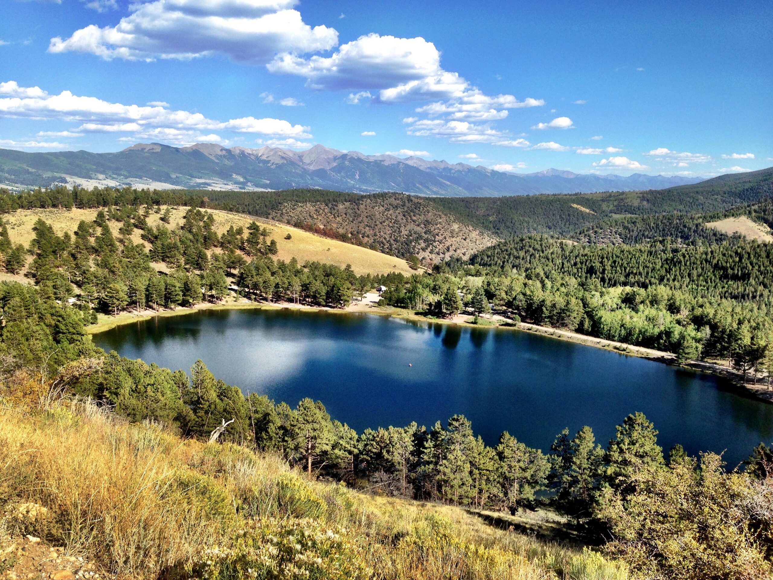 A scenic view of a serene lake surrounded by lush greenery and rolling hills, under a blue sky with fluffy white clouds. The landscape features a mix of trees and grassy areas, with distant mountains visible in the background. Marshall Pass Road / #200 / #203 / #243 mountain bike trail.