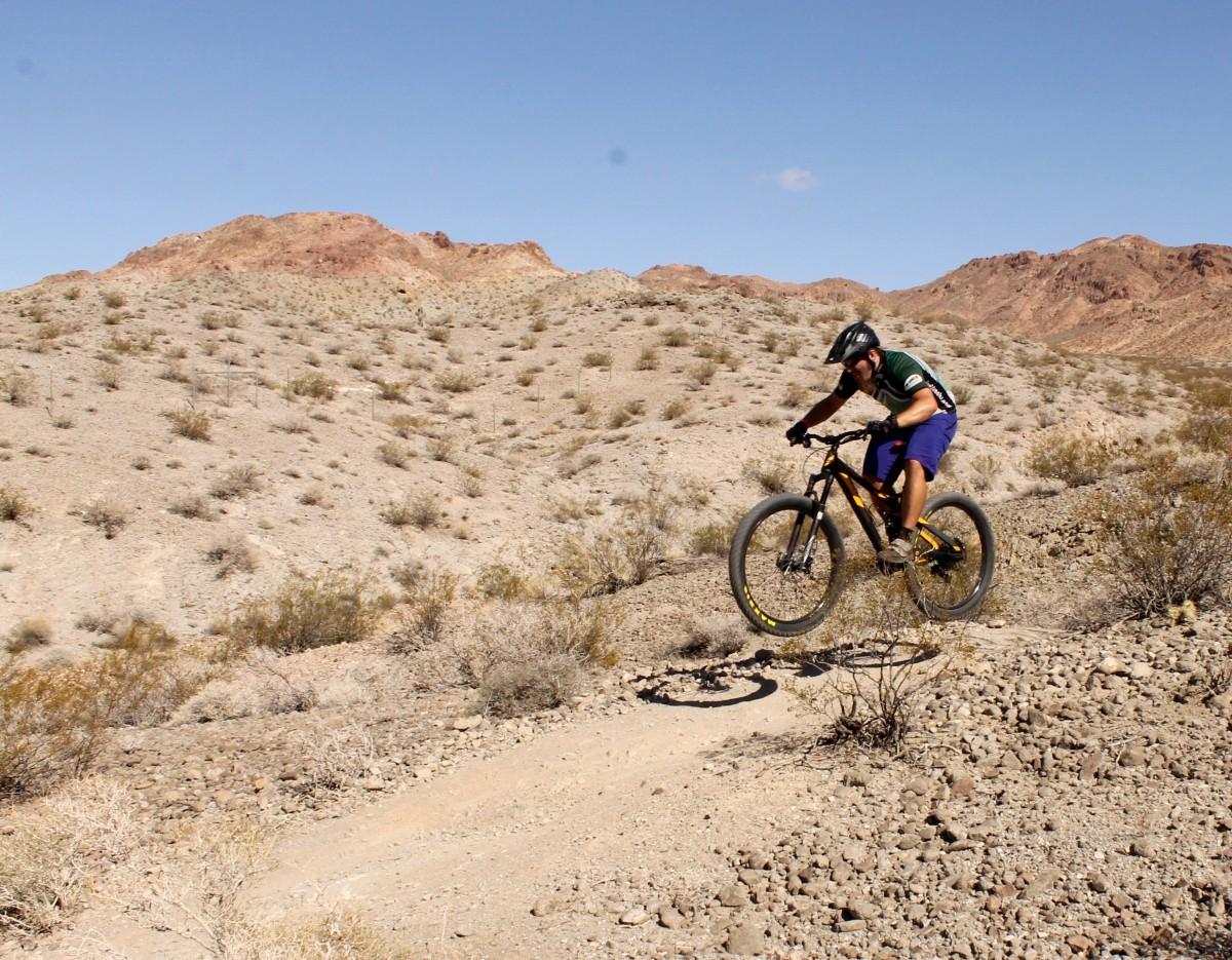 A mountain biker in mid-air performing a jump on a rocky trail in a desert landscape, surrounded by sparse vegetation and rugged hills under a clear blue sky. Bootleg Canyon mountain bike trail.
