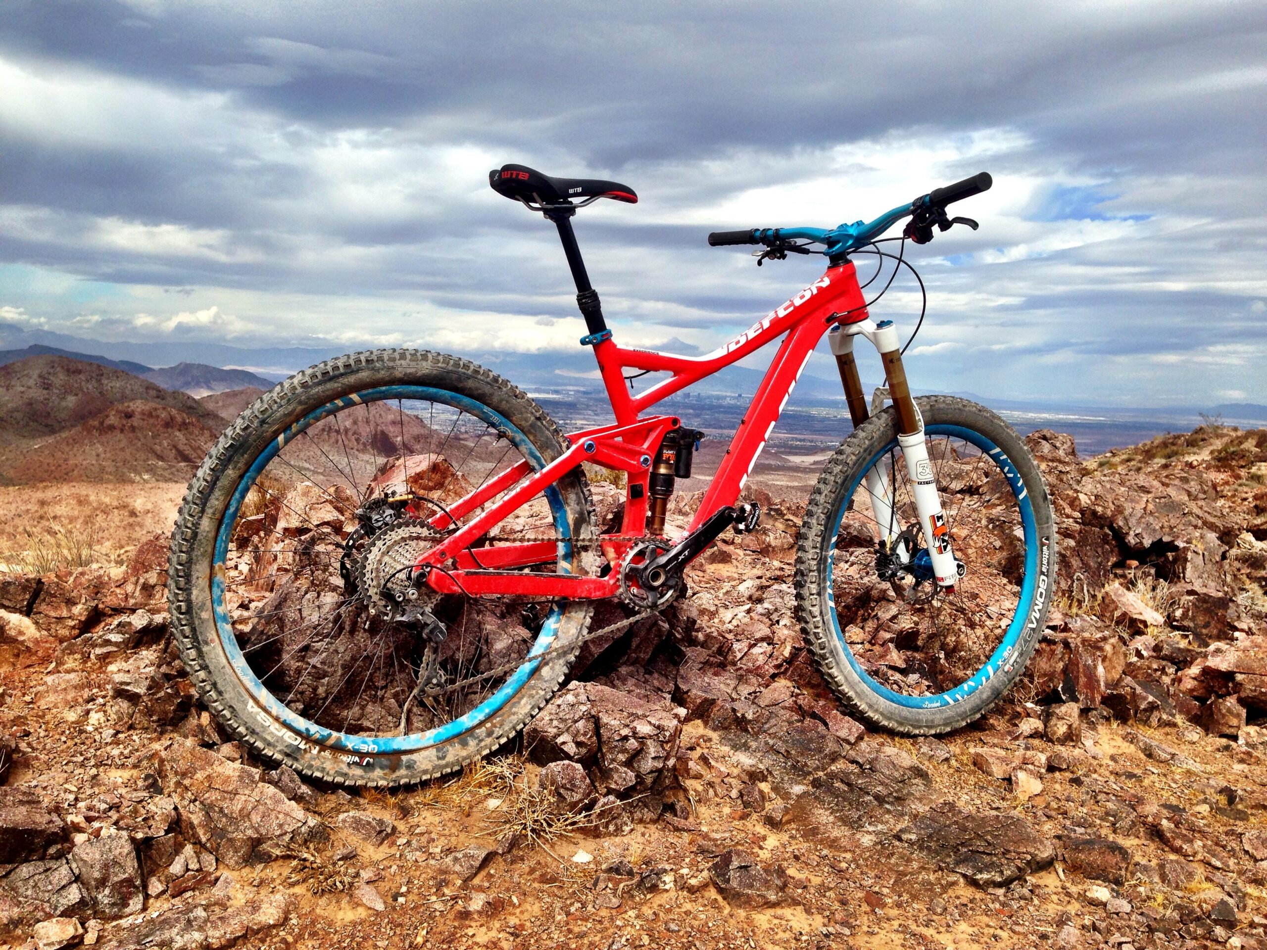 A red mountain bike positioned on rocky terrain with distant mountains and a cloudy sky in the background. The bike features blue accents on the wheels and components, highlighting its design against the natural landscape. Bootleg Canyon mountain bike trail.