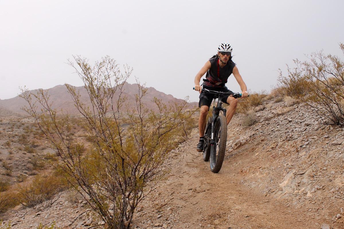A cyclist riding a mountain bike down a rocky trail in a desert landscape, with shrubs and mountains in the background. The rider is wearing a helmet and athletic gear, focused on navigating the uneven terrain. The sky is overcast, creating a muted atmosphere. Bootleg Canyon mountain bike trail.