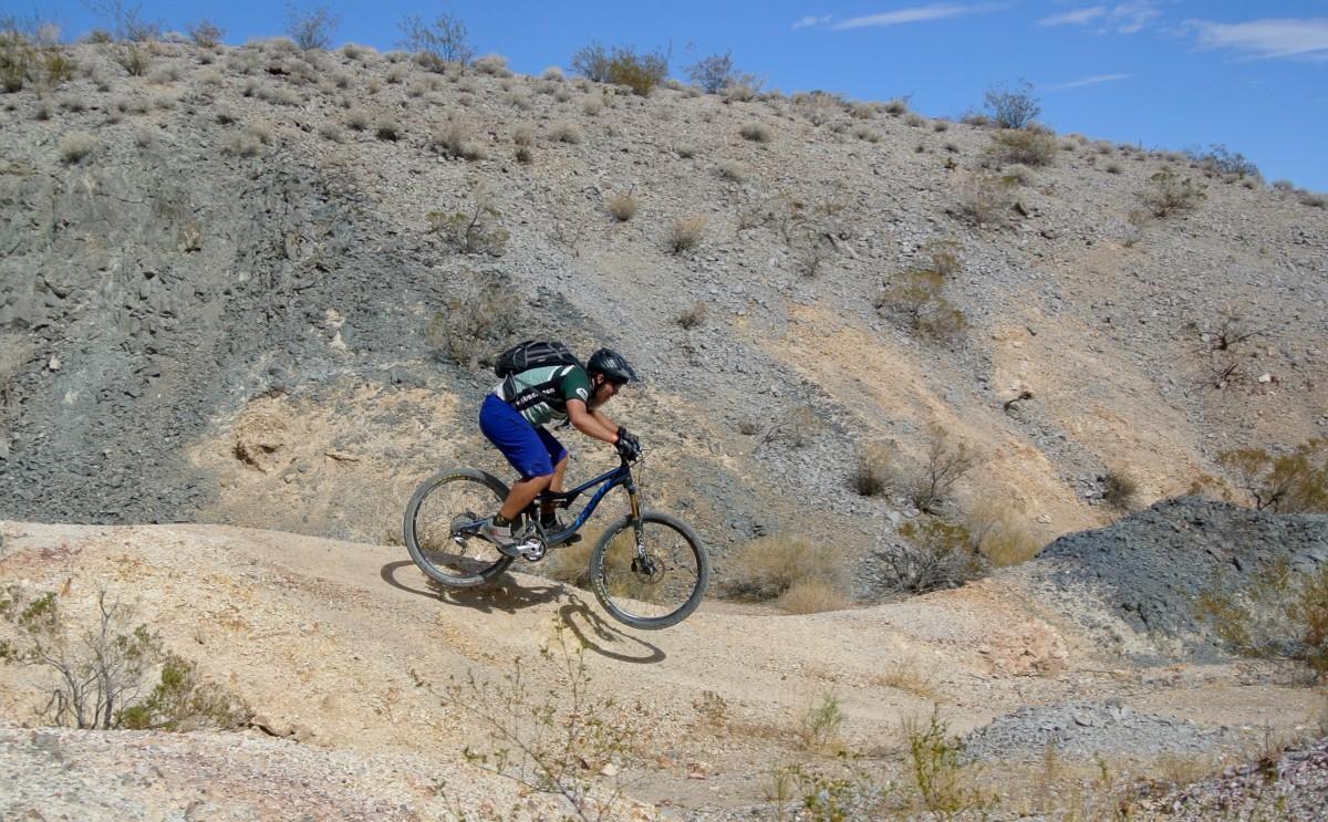 A mountain biker navigating a rocky trail, captured mid-jump with a backdrop of dry, rugged terrain and sparse vegetation under a blue sky. Bootleg Canyon mountain bike trail.