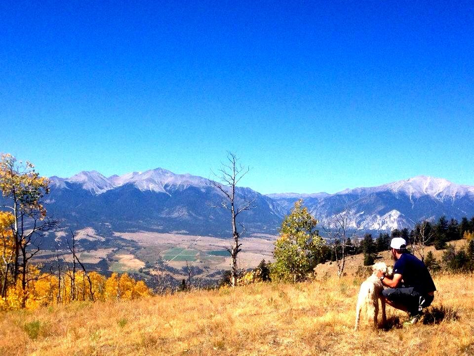 A person kneels beside a dog on a grassy hillside, overlooking a vast landscape of mountains and valleys under a clear blue sky. The scene features autumn foliage in the foreground and snow-capped peaks in the background, creating a serene outdoor atmosphere. Aspen Ridge / Road #185 mountain bike trail.
