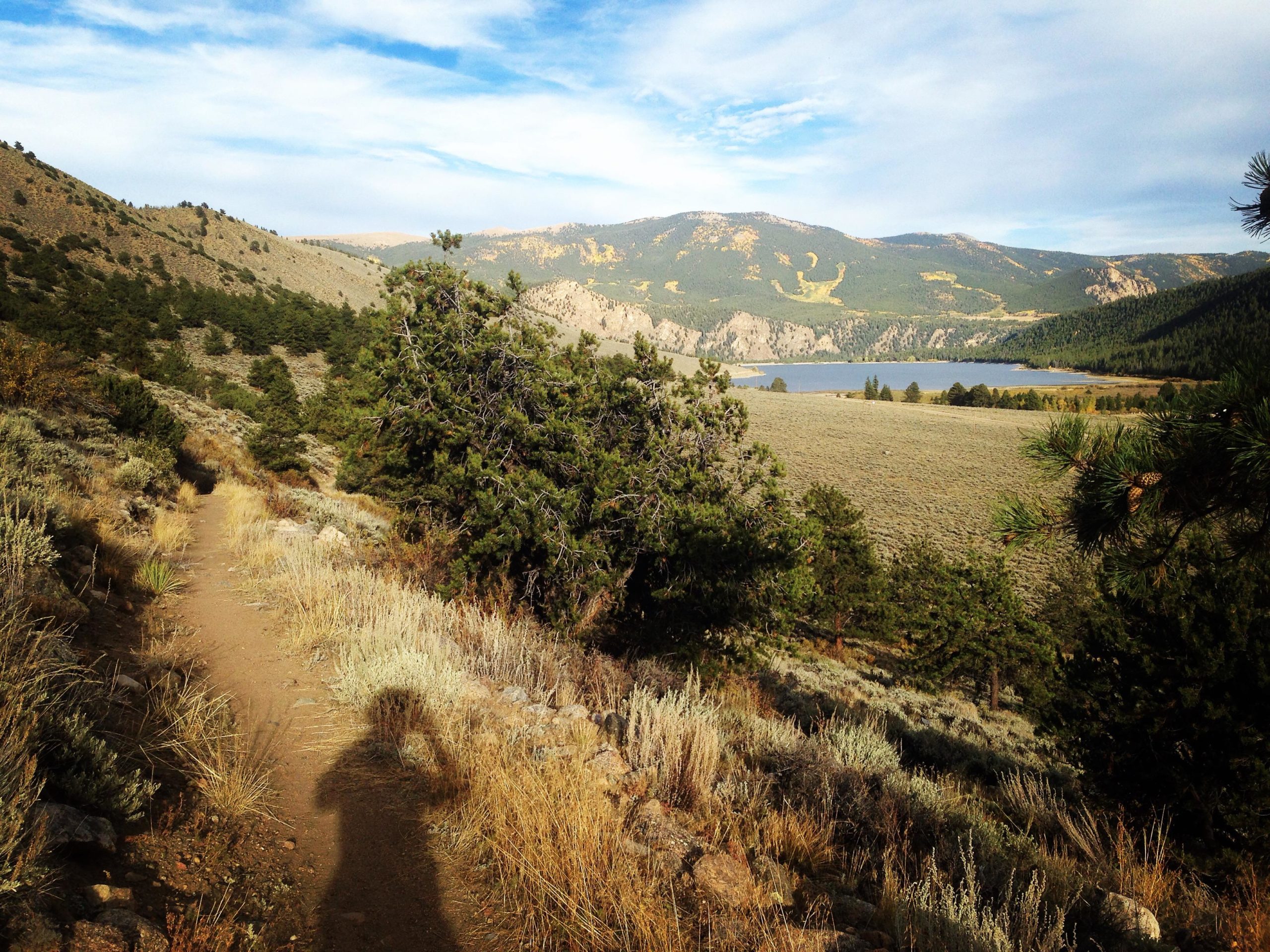 A scenic landscape featuring a dirt hiking trail winding through golden grasses and shrubs, bordered by trees. In the background, rolling hills lead to a serene lake surrounded by mountains under a partly cloudy sky. A shadow of the photographer is visible on the path. Colorado Trail: Clear Creek Thd to Lake View CG / Hwy 82 mountain bike trail.