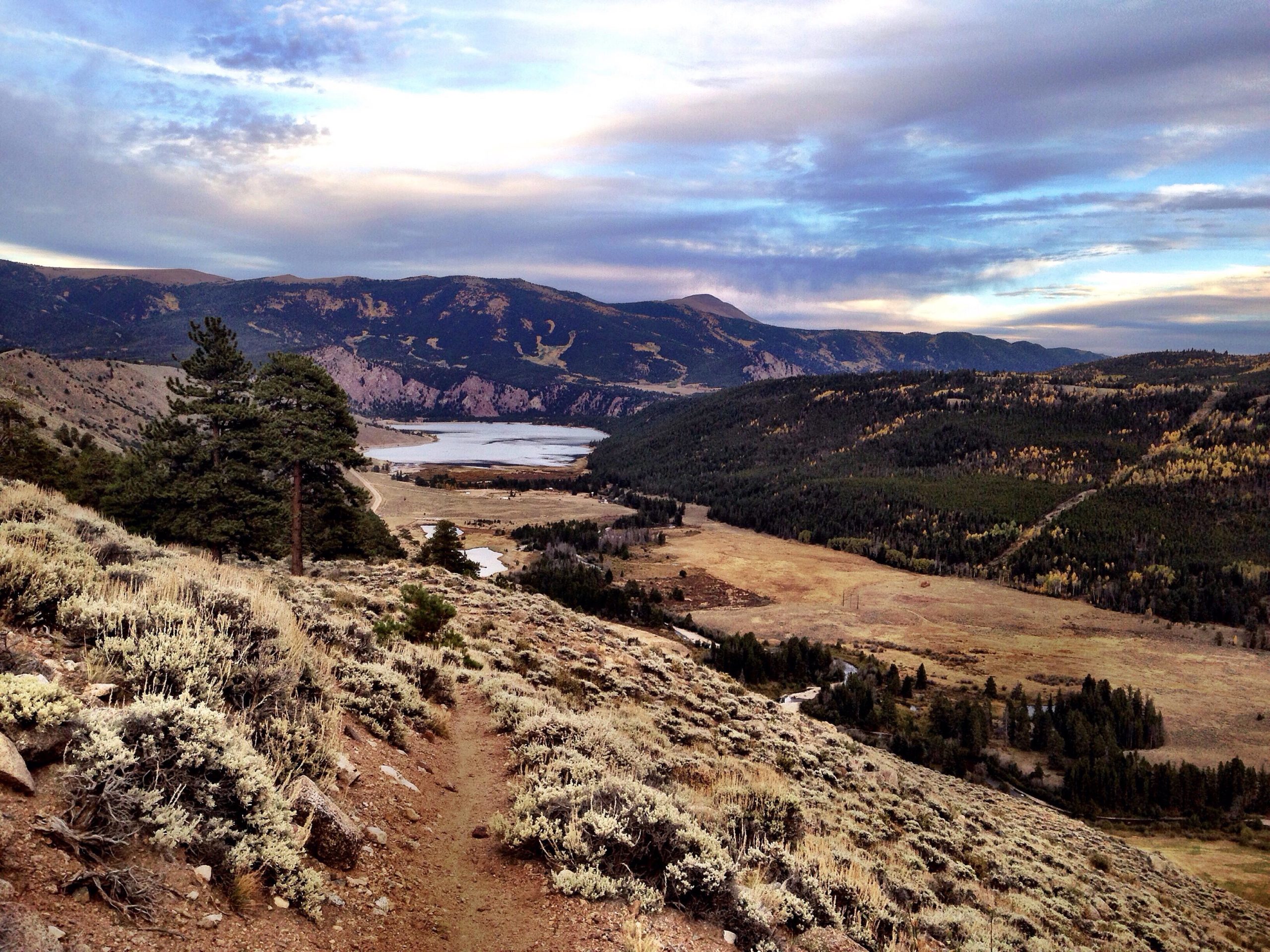 A scenic view from a hillside overlooking a valley, with a winding river and lake in the foreground. Pine trees and shrubs are scattered throughout the terrain, while distant mountains provide a dramatic backdrop under a partially cloudy sky. Colorado Trail: Clear Creek Thd to Lake View CG / Hwy 82 mountain bike trail.