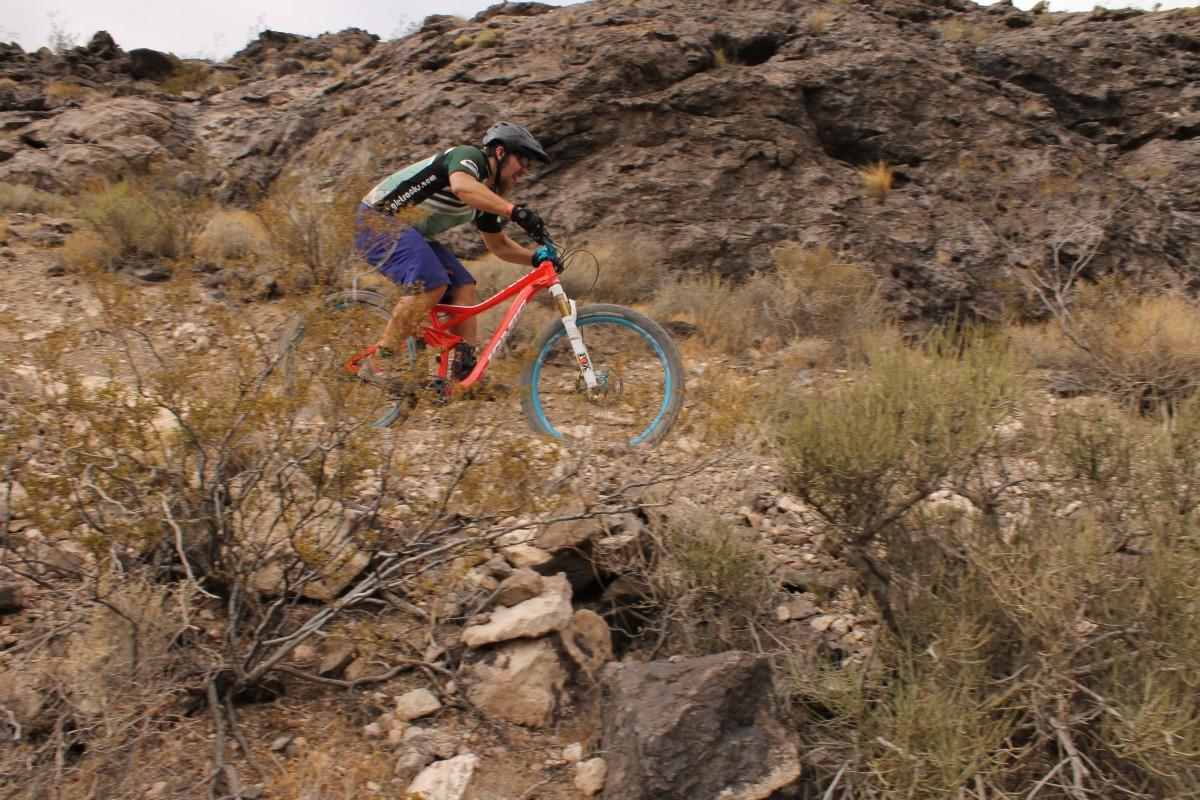 A mountain biker wearing a helmet and protective gear rides over rocky terrain, navigating a trail surrounded by sparse vegetation and boulders. The bike has distinctive blue rims and the rider is in an athletic posture, emphasizing motion and speed. Bootleg Canyon mountain bike trail.