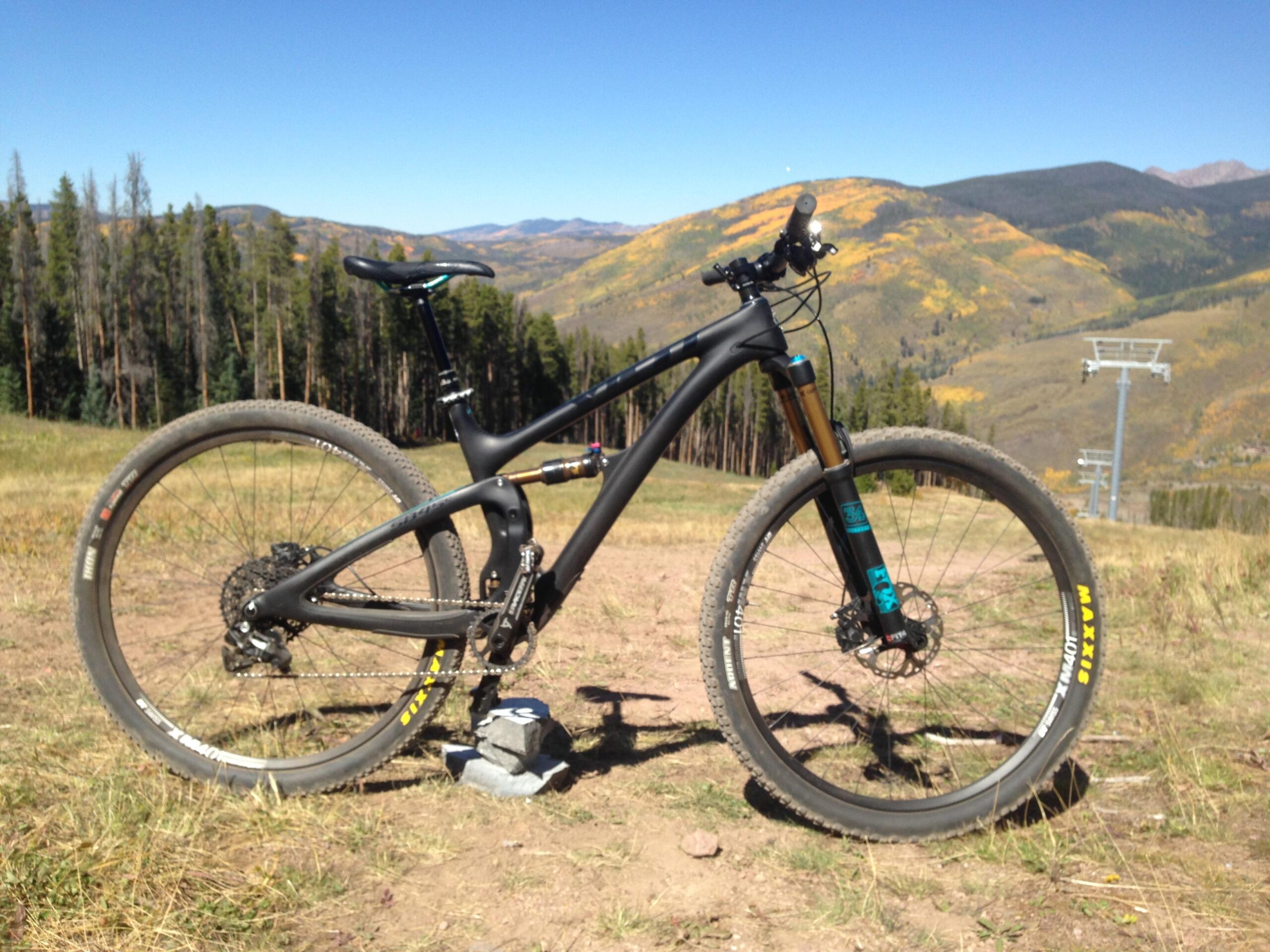 A black mountain bike with thick tires, positioned on a dirt trail with a scenic backdrop of mountains and trees. The sky is clear and blue, showcasing a vibrant landscape with hints of autumn foliage on the hillsides. A ski lift can be seen in the distance. Vail Mountain Bike Park mountain bike trail.