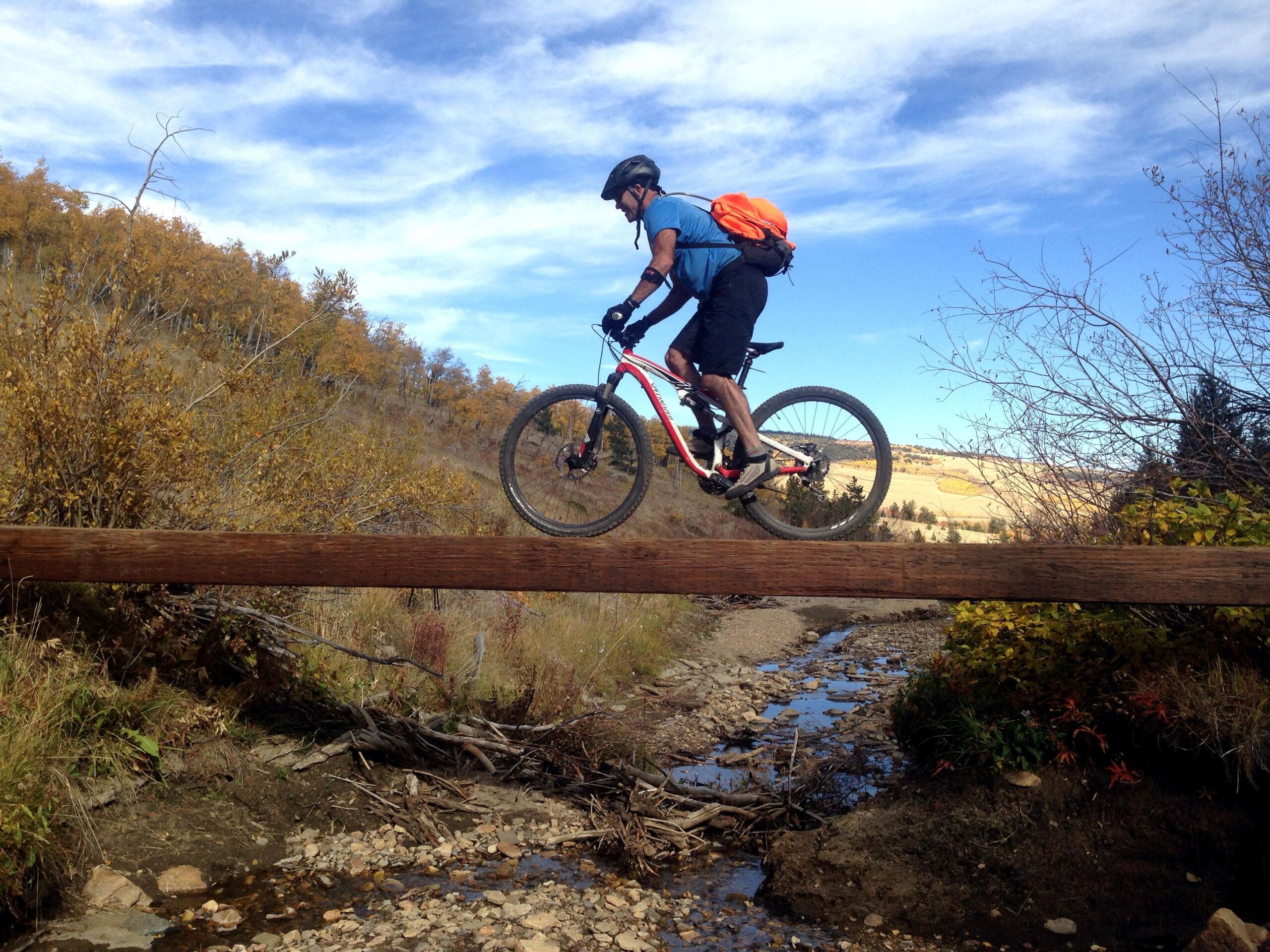 A mountain biker riding across a narrow wooden bridge over a shallow stream, surrounded by autumn foliage and a clear blue sky. Sheep Creek mountain bike trail.