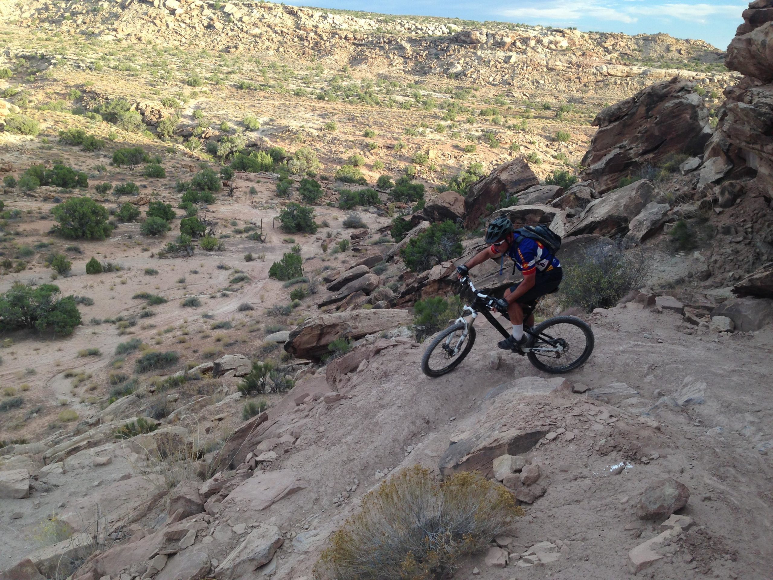 A mountain biker riding down a rocky trail in a desert landscape with sparse vegetation and distant hills in the background. The biker is wearing a helmet and colorful cycling gear, navigating the uneven terrain. Sovereign Single Track mountain bike trail.