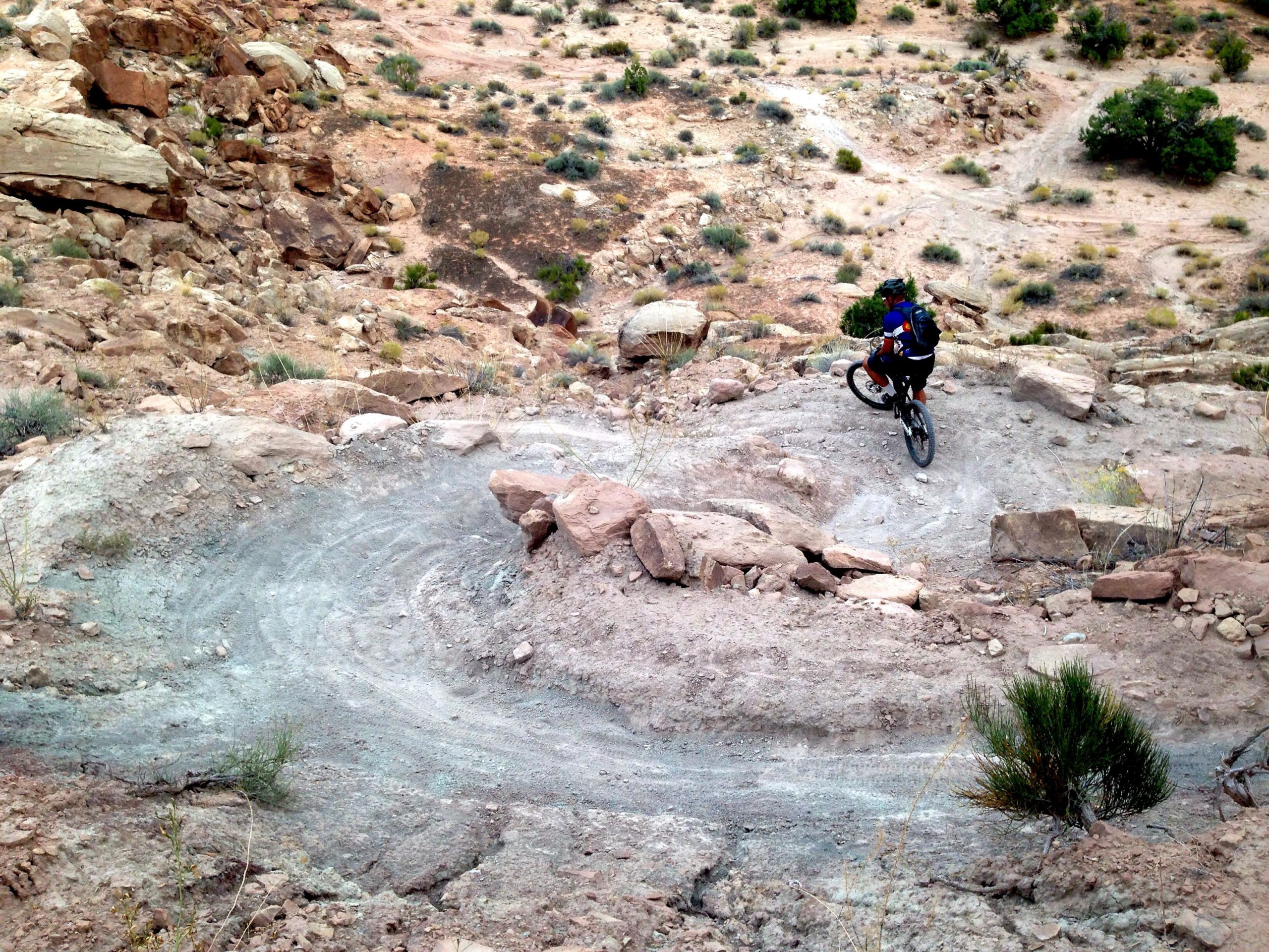 A mountain biker navigates a rocky trail on a rugged landscape, featuring winding paths and scattered vegetation. The terrain includes loose rocks and dirt, highlighting the challenges of off-road biking. Sovereign Single Track mountain bike trail.
