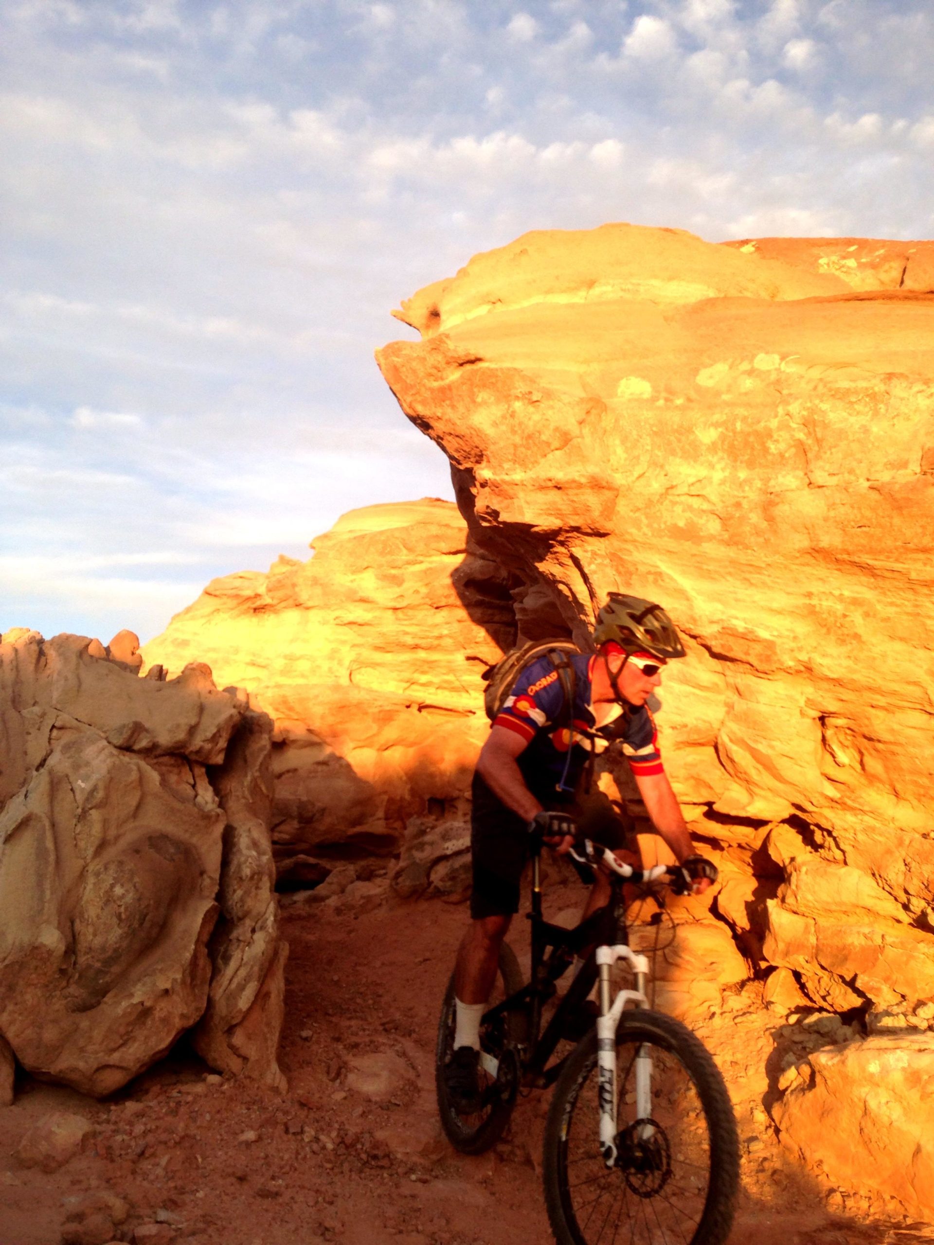 A mountain biker navigating a rocky trail surrounded by reddish-orange rock formations during sunset. The biker is wearing a helmet and cycling gear, and the sun casts warm light on the landscape and the rider. Sovereign Single Track mountain bike trail.