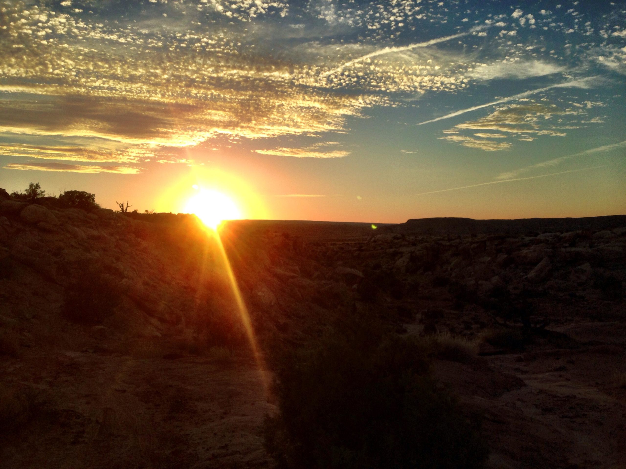 A stunning sunset over a desert landscape, with the sun low on the horizon casting warm golden light and creating a flare of sunlight. Fluffy clouds partially cover the sky, and the rocky terrain is dotted with sparse vegetation, enhancing the serene beauty of the scene. Sovereign Single Track mountain bike trail.